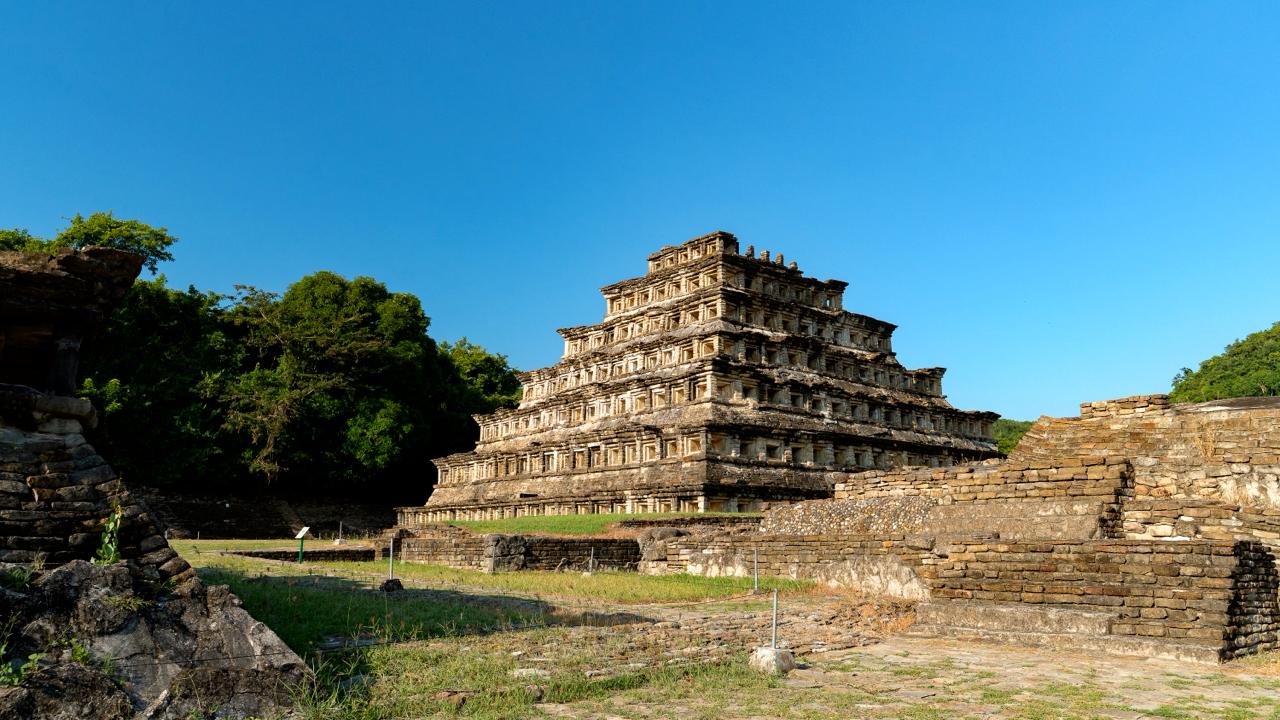 Fotografía A Cielo Abierto Que Muestra En Un Plano General La Pirámide De Los Nichos En La Zona Arqueológica De El Tajín. La Estructura Piramidal Está Compuesta Por Múltiples Niveles Escalonados Construidos En Piedra Labrada, Destacando Las Filas Simétricas De Nichos Cuadrangulares En Cada Una De Sus Caras. A La Izquierda De La Pirámide Se Observa Una Densa Vegetación De Árboles Verdes, Mientras Que En Primer Plano Se Aprecian Restos De Muros Y Estructuras Arqueológicas Menores Rodeadas De Pasto Corto. El Cielo Es Completamente Despejado Y De Un Color Azul Intenso, Iluminando La Escena Con Luz Solar Directa.