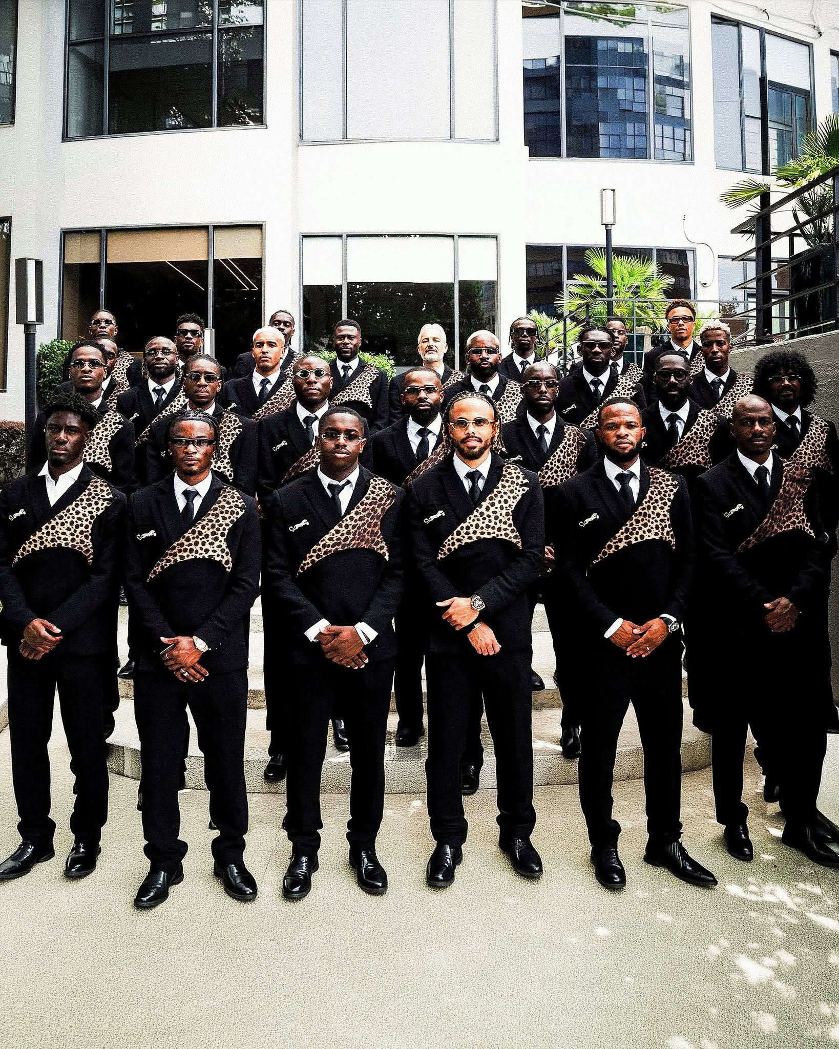 El regreso (con mucho estilo) de la república democrática del congo al mundial 2026 la revista del sureste mx Fotografía Grupal De La Delegación Completa De La Selección De Futbol De La República Democrática Del Congo Posando En Unas Escaleras Exteriores Frente A Un Edificio Blanco. Todos Los Integrantes Visten Trajes Sastre Negros Idénticos Con Una Banda Cruzada De Estampado De Leopardo En El Pecho, Camisas Blancas, Corbatas Negras Y Gafas De Sol, Mostrando Una Imagen Unificada Y Elegante.