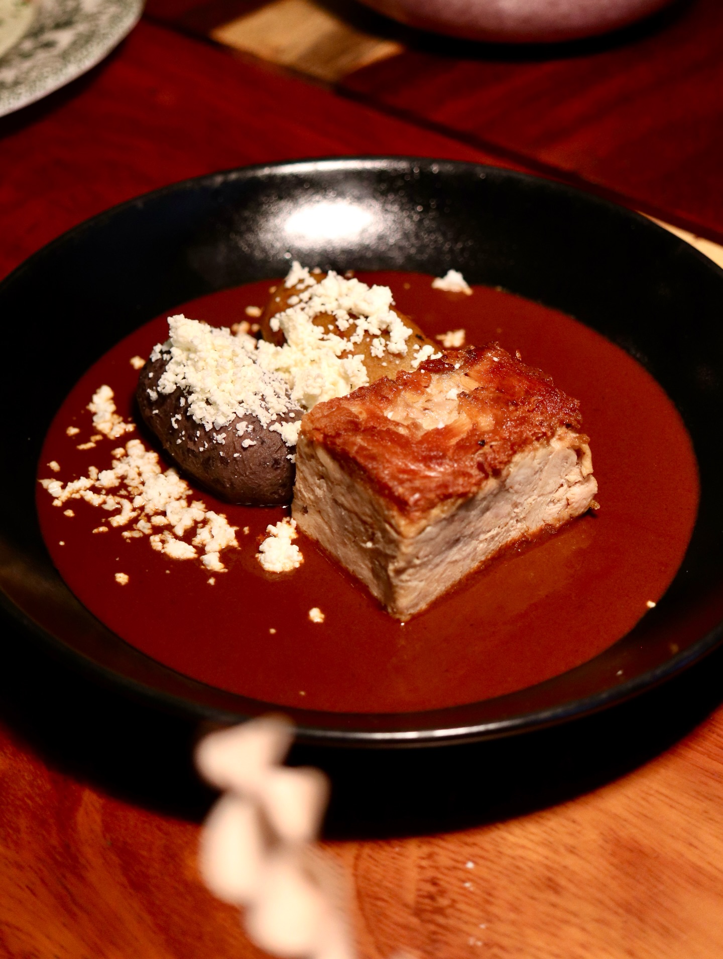 Excelencia gastronómica en villahermosa: 3 restaurantes imperdibles la revista del sureste mx Una Vista De Primer Plano De Un Plato De Autor Servido En Un Tazón Negro, Que Presenta Una Porción Rectangular Y Dorada De Lechón Crujiente Sumergida En Una Salsa De Mole De Color Rojo Oscuro Y Rico. Junto A Él Hay Un Platanito Relleno Y Una Marqueta De Frijoles, Ambos Cilíndricos Y Cubiertos Generosamente Con Queso Fresco Desmoronado, Sobre Una Base De Mole Adicional Y Guarniciones De Queso.