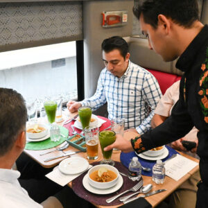 Sabores sobre rieles: el tren maya consolida la experiencia de su tren restaurante "janal" la revista del sureste mx Fotografía En Perspectiva Diagonal Tomada Dentro Del Comedor Del Tren Maya Durante Una Prueba De Servicio. Sentados A Una Mesa De Madera Clara Junto A La Ventana, Cuatro Comensales Del Sector Turístico E Institucional Observan Los Platillos Mientras Un Mesero, Vestido Con Una Camisa Negra Decorada Con Bordados Florales Tradicionales De Color Naranja En Los Brazos Y El Pecho, Sirve Un Vaso Con Una Bebida Verde.