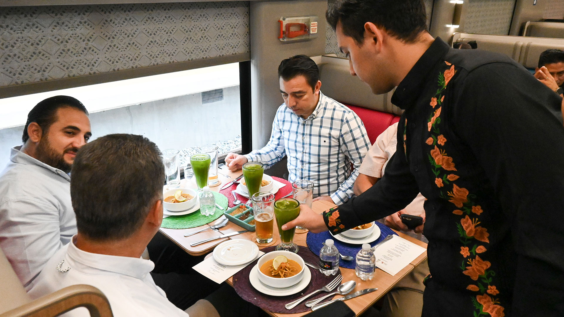 Fotografía En Perspectiva Diagonal Tomada Dentro Del Comedor Del Tren Maya Durante Una Prueba De Servicio. Sentados A Una Mesa De Madera Clara Junto A La Ventana, Cuatro Comensales Del Sector Turístico E Institucional Observan Los Platillos Mientras Un Mesero, Vestido Con Una Camisa Negra Decorada Con Bordados Florales Tradicionales De Color Naranja En Los Brazos Y El Pecho, Sirve Un Vaso Con Una Bebida Verde.