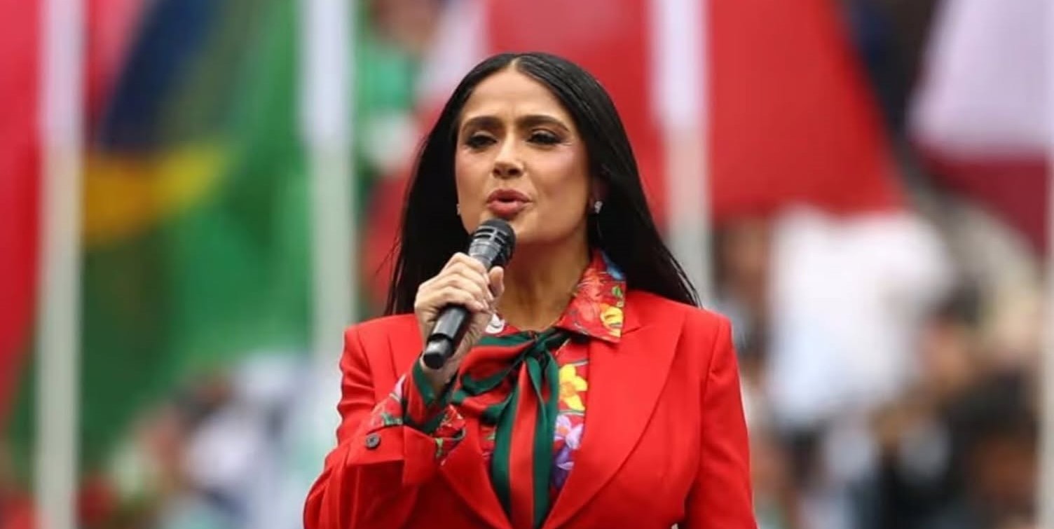 Una Fotografía De Plano Medio Que Captura A La Actriz Salma Hayek Dirigiendo Unas Palabras Al Público Durante La Inauguración De La Copa Del Mundo De La Fifa México 2026 En El Estadio Ciudad De México. Viste Un Traje Sastre En Color Rojo Intenso, Complementado Con Una Blusa Estampada De Flores Multicolores Y Una Corbata De Lazo Con Franjas Verde Y Rojo, De La Firma Gucci.