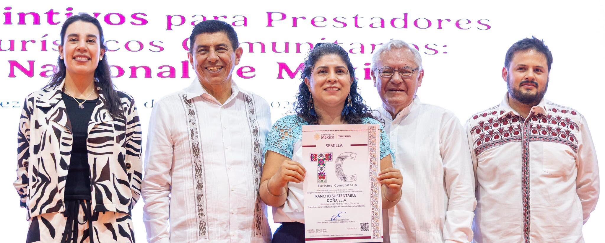 Cinco Personas Posan De Pie En Un Escenario Durante Un Evento Oficial. En El Centro, Una Mujer Sonriente Sostiene Un Reconocimiento Impreso De La Secretaría De Turismo Del Gobierno De México Con El Logotipo &Quot;Tc Turismo Comunitario&Quot; Y El Nombre &Quot;Rancho Sustentable Doña Elia&Quot;. A Su Izquierda Se Encuentran La Secretaria De Turismo, Josefina Rodríguez Zamora, Vistiendo Un Conjunto Estampado En Blanco Y Negro, Y El Gobernador De Oaxaca, Salomón Jara Cruz, Con Una Guayabera Blanca Tradicional. A La Derecha De La Galardonada, Completan El Grupo Dos Funcionarios Públicos Masculinos Con Vestimenta Formal Y Artesanal. Al Fondo Se Observa Una Pantalla Con Letras En Color Rosa.