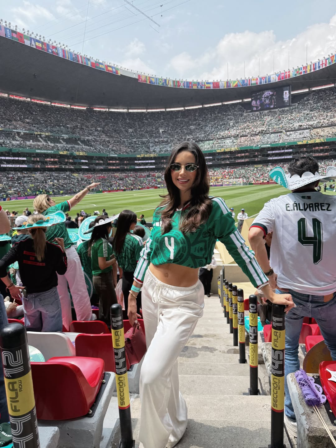 Plano Medio Entero De La Modelo Sofía Toache Posando Sonriente En Las Escaleras De Las Tribunas De Un Estadio De Fútbol Completamente Lleno Durante El Mundial 2026. Viste Un Jersey Verde De La Selección Mexicana Estilo Crop Top Con El Número 4 En El Pecho, Pants Blancos Holgados Y Lentes De Sol. En Su Mano Derecha Sostiene Un Bolso Color Vino Mientras Se Apoya En El Barandal. A Su Lado Derecho Destaca Un Aficionado De Espaldas Portando Una Playera Blanca Con El Dorsal Número 4 Y El Apellido &Quot;E. Álvarez&Quot;.