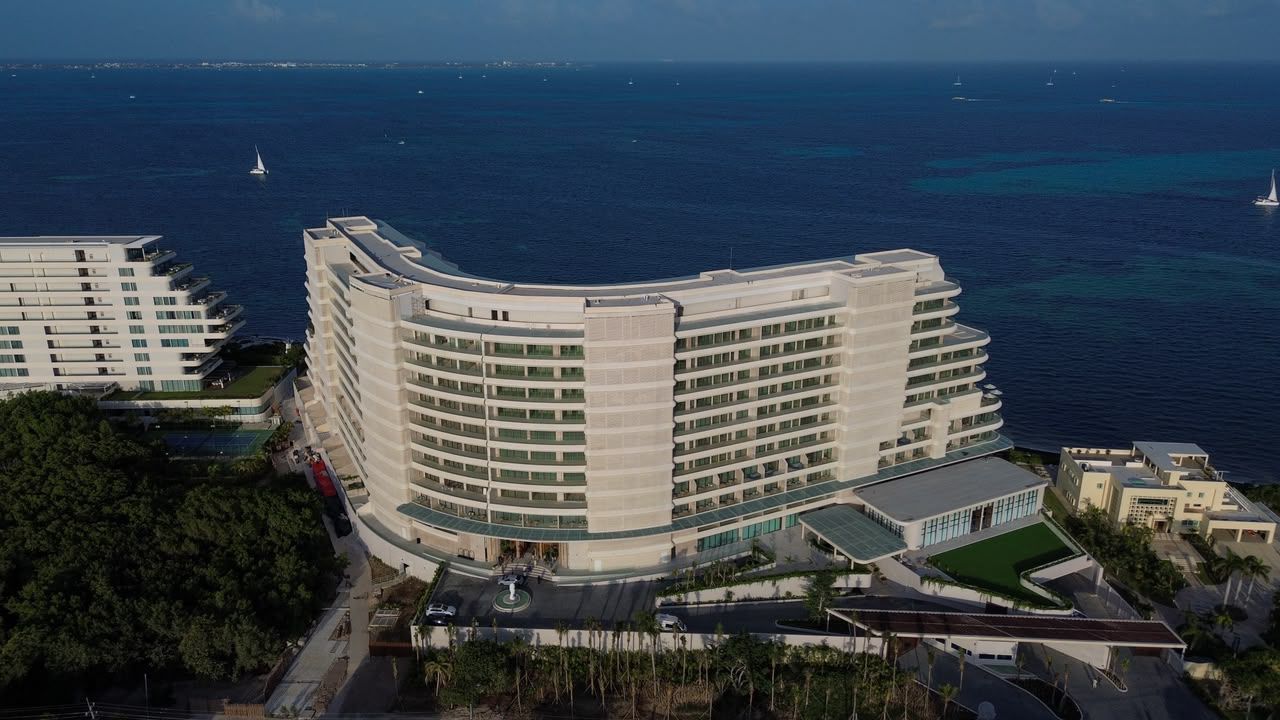 Una Toma Aérea De Un Moderno Hotel St. Regis Costa Mujeres En La Costa. El Hotel Tiene Una Forma Curva Única Y Está Situado Junto A Un Denso Bosque Tropical. Varias Canchas De Tenis Y Una Gran Piscina Están En El Lugar.