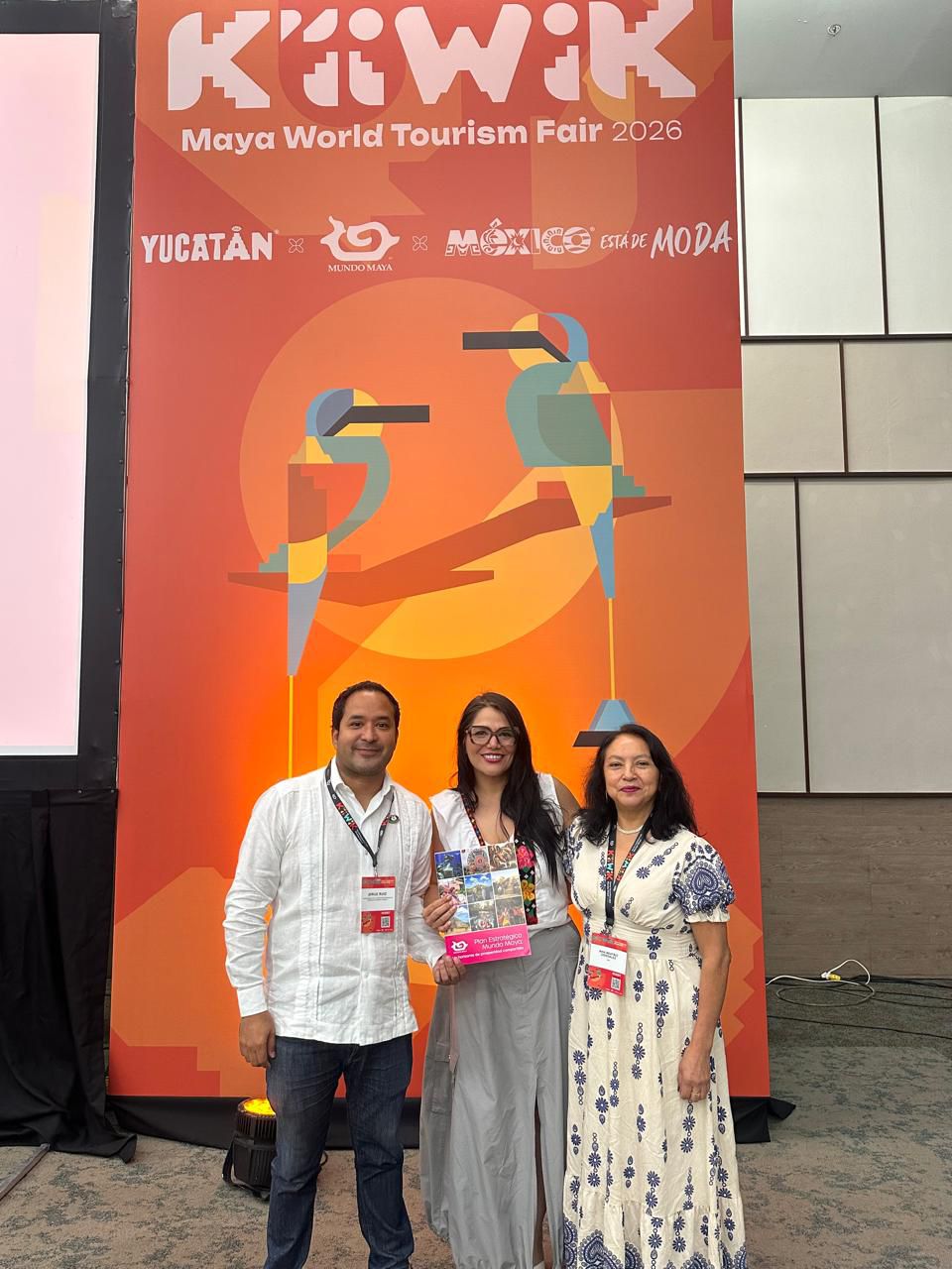 Un Hombre Y Dos Mujeres Posan De Pie Y Sonrientes Frente A Una Gran Mampara Promocional De Color Naranja Brillante En Un Salón De Eventos. El Panel Del Fondo Muestra En La Parte Superior El Logotipo &Quot;K’íiwik Maya World Tourism Fair 2026&Quot;, Seguido De Los Distintivos De &Quot;Yucatán&Quot;, &Quot;Mundo Maya&Quot; Y &Quot;México Está De Moda&Quot;; Al Centro Del Diseño, Destaca Una Ilustración Geométrica De Dos Pájaros Toh Posados Sobre Ramas Frente A Un Círculo Solar Amarillo.