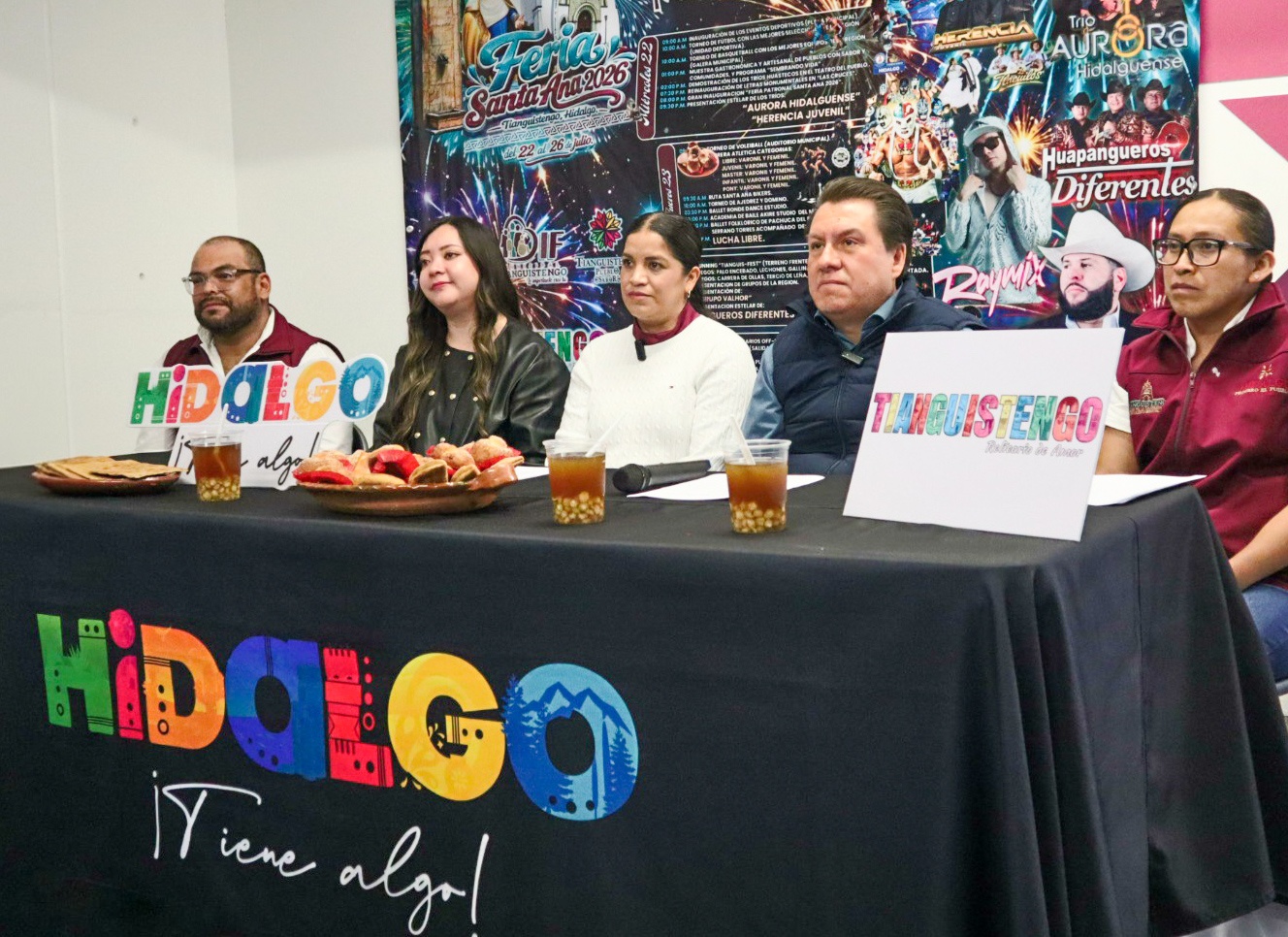 Fotografía Tomada Desde Un Ángulo Diagonal Que Muestra A Cinco Funcionarios Públicos Sentados En Una Mesa De Presídium Cubierta Con Un Mantel Negro. En El Frente Del Mantel Destaca En Letras Grandes Y Multicolores El Logotipo Turístico &Quot;Hidalgo ¡Tiene Algo!&Quot;. Sobre La Mesa Hay Vasos De Plástico Con La Bebida Tradicional Axocotl, Un Platón Con Pan De Dulce Artesanal, Un Micrófono Y Un Letrero Con La Palabra &Quot;Tianguistengo&Quot;. Detrás De Los Asistentes Se Aprecia Una Gran Lona Promocional De La Feria Patronal Con Imágenes De Artistas De Música Regional, Luchadores Y Detalles Del Programa Del 22 Al 26 De Julio.
