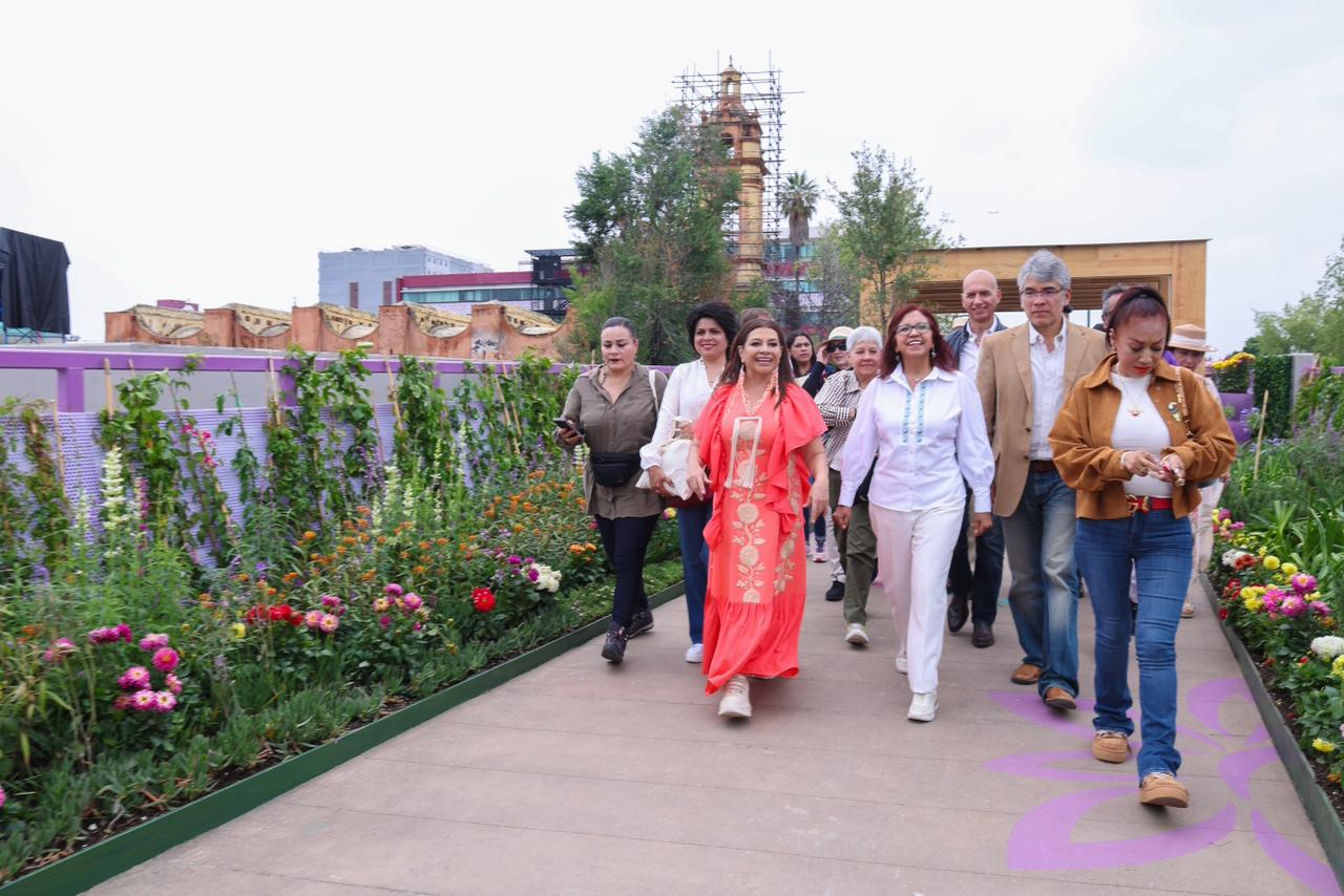 Tlallipan: cdmx inaugura el primer segundo piso peatonal y jardín flotante del continente la revista del sureste mx Una Fotografía De Plano General Captura A Un Grupo Numeroso De Personas, Predominantemente Mujeres Adultas, Caminando Alegremente Hacia La Cámara Por Un Andador Elevado Y Arbolado. El Camino Es De Concreto Con Patrones De Flores Púrpuras Integrados En El Suelo, Flanqueado A Ambos Lados Por Exuberantes Jardines De Flores Vibrantes (Rosas, Rojas, Naranjas, Verdes) Y Camas Altas De Plantación Con Vallas De Color Púrpura Claro Cubiertas De Vegetación Trepadora.