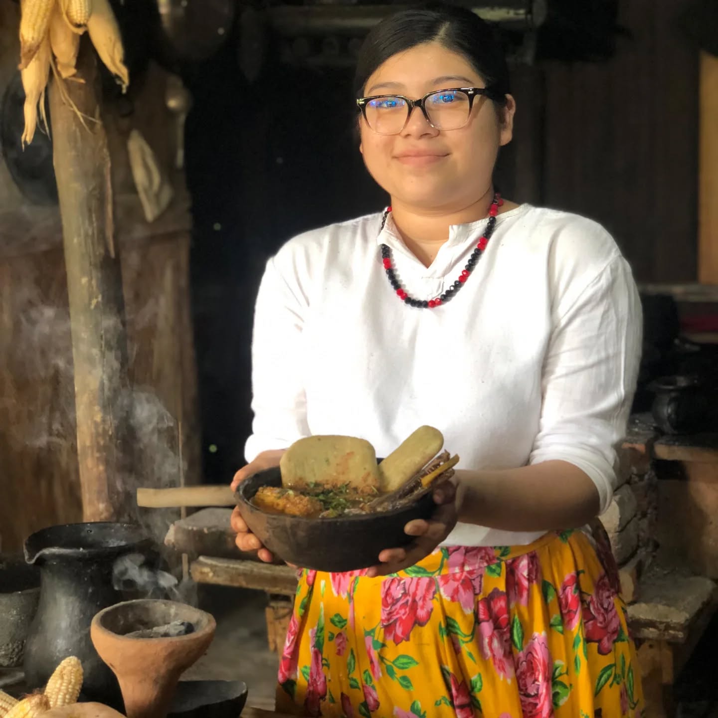El edén de los sabores: tres paradas gastronómicas imperdibles para redescubrir el tabasco ancestral y tradicional la revista del sureste mx Una Mujer Joven Con Anteojos, Blusa Blanca, Collar De Cuentas Rojas Y Negras, Y Una Falda Larga Amarilla Con Un Estampado Floral Rosa, Sonríe Mientras Sostiene Con Ambas Manos Un Plato Hondo De Barro Oscuro. El Plato Contiene Un Guiso Caldoso Decorado Con Hierbas Frescas Y Acompañado De Gruesas Tortillas De Maíz Tradicionales. Al Fondo, En Una Cocina Rústica De Humo, Se Aprecian Elementos Tradicionales Como Mazorcas De Maíz Colgadas, Un Fogón De Leña Y Utensilios De Barro De Los Que Emana Un Ligero Vapor.