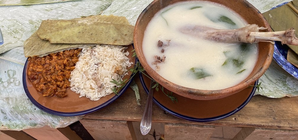 Un Plato De Uliche Tradicional De Tabasco Servido En Un Cajete De Barro Con Una Pieza De Pavo Y Caldo Blanco Espeso Con Hojas Verdes. Al Lado, Un Plato De Barro Con Arroz Blanco, Guiso De Carne En Salsa Roja Y Tortillas De Maíz Nuevo Dobladas, Todo Dispuesto Sobre Una Mesa Rústica De Madera Con Hojas De Plátano De Fondo.