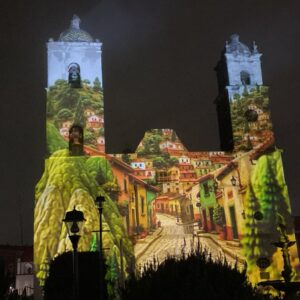 Videomapping en real del monte: hidalgo impulsa turismo inmersivo por el mundial 2026 la revista del sureste mx Fotografía Nocturna De La Fachada Barroca Y Las Dos Torres De La Parroquia De Nuestra Señora De La Asunción En Real Del Monte, Hidalgo. Toda La Estructura Del Templo Sirve Como Pantalla Para Una Colorida Proyección De Videomapping Que Recrea Una Calle Empedrada, Flanqueada Por Casas Coloniales De Tonos Amarillos, Verdes Y Naranjas, Que Ascienden Hacia Colinas Boscosas. En Primer Plano Y En Silueta Oscura, Se Aprecian Faroles Públicos Y Arbustos De La Plaza Principal.