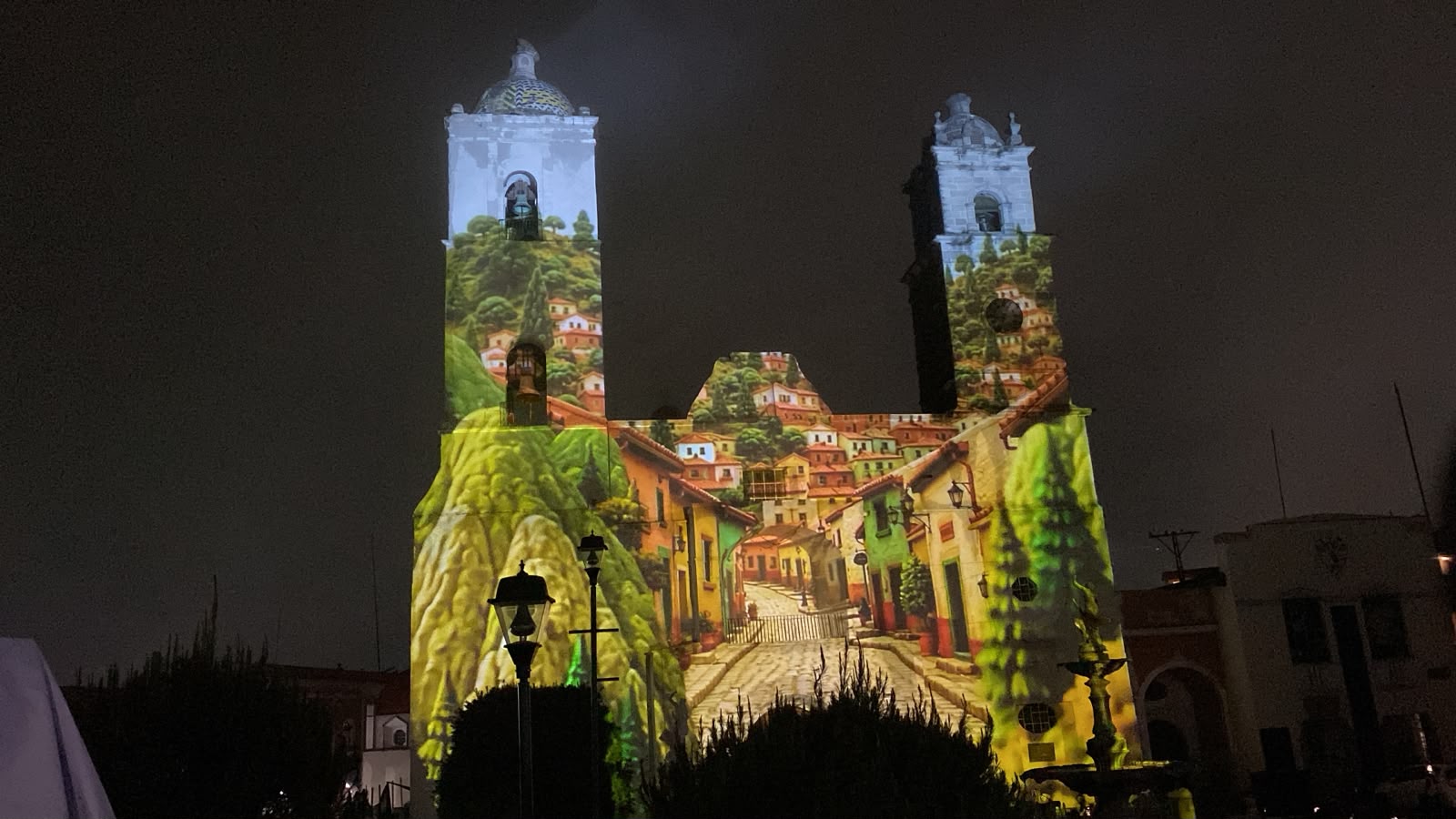 Videomapping en real del monte: hidalgo impulsa turismo inmersivo por el mundial 2026 la revista del sureste mx Fotografía Nocturna De La Fachada Barroca Y Las Dos Torres De La Parroquia De Nuestra Señora De La Asunción En Real Del Monte, Hidalgo. Toda La Estructura Del Templo Sirve Como Pantalla Para Una Colorida Proyección De Videomapping Que Recrea Una Calle Empedrada, Flanqueada Por Casas Coloniales De Tonos Amarillos, Verdes Y Naranjas, Que Ascienden Hacia Colinas Boscosas. En Primer Plano Y En Silueta Oscura, Se Aprecian Faroles Públicos Y Arbustos De La Plaza Principal.