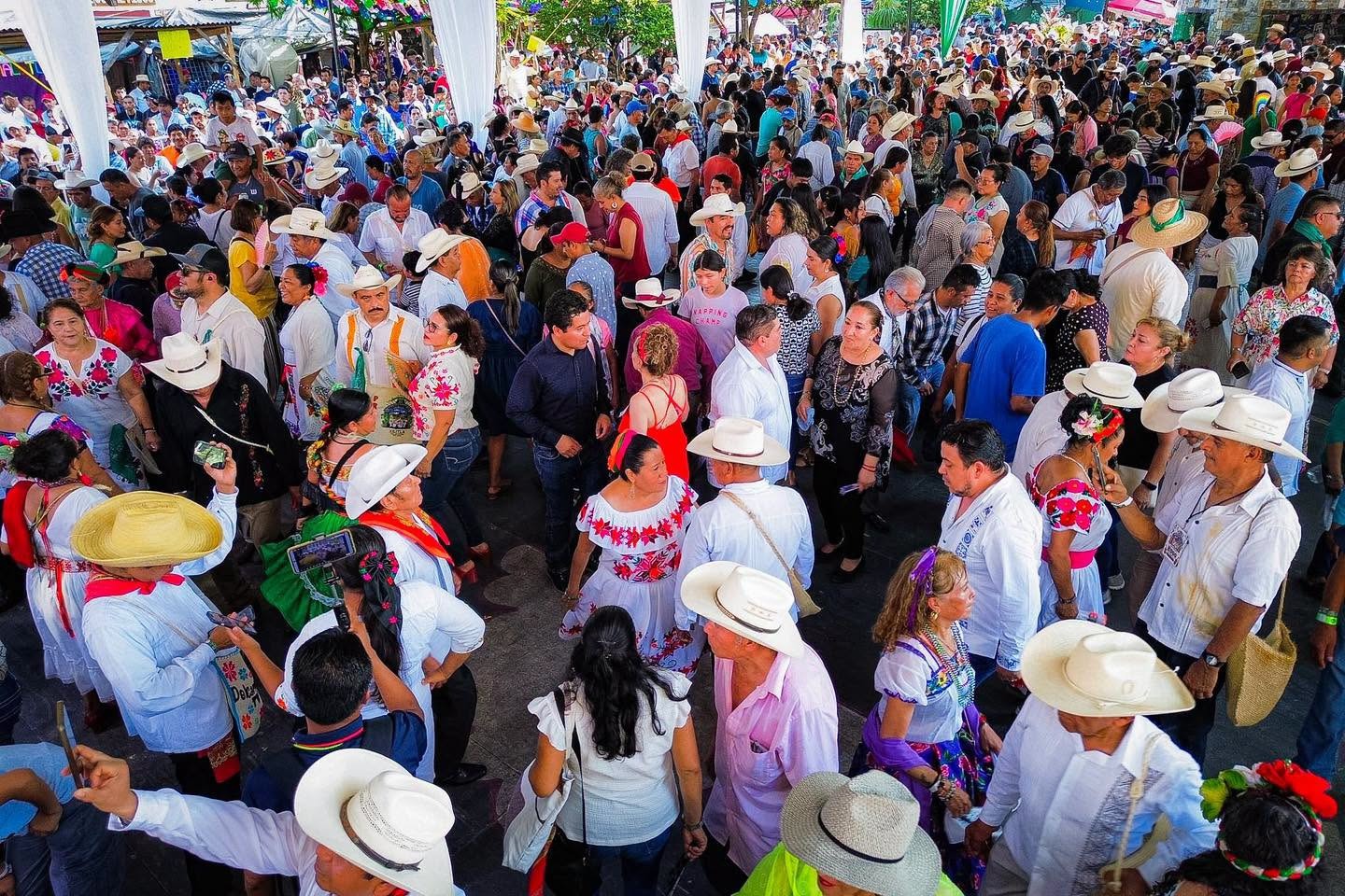 Una Toma Aérea En Plano General Que Muestra A Una Enorme Multitud De Personas Congregadas En Una Plaza Pública Al Aire Libre. La Mayoría Viste Indumentaria Tradicional De Huapango: Las Mujeres Portan Blusas Blancas Con Coloridos Bordados Florales Y Faldas Folclóricas, Mientras Que Los Hombres Llevan Guayaberas O Camisas Blancas, Pantalones Y Sombreros Vaqueros De Color Claro. Se Observa A La Gente Distribuida Por Parejas Y En Grupos Sobre El Pavimento, Interactuando Y Preparándose Para El Baile Colectivo En Un Ambiente Festivo Y Culturalmente Vibrante.
