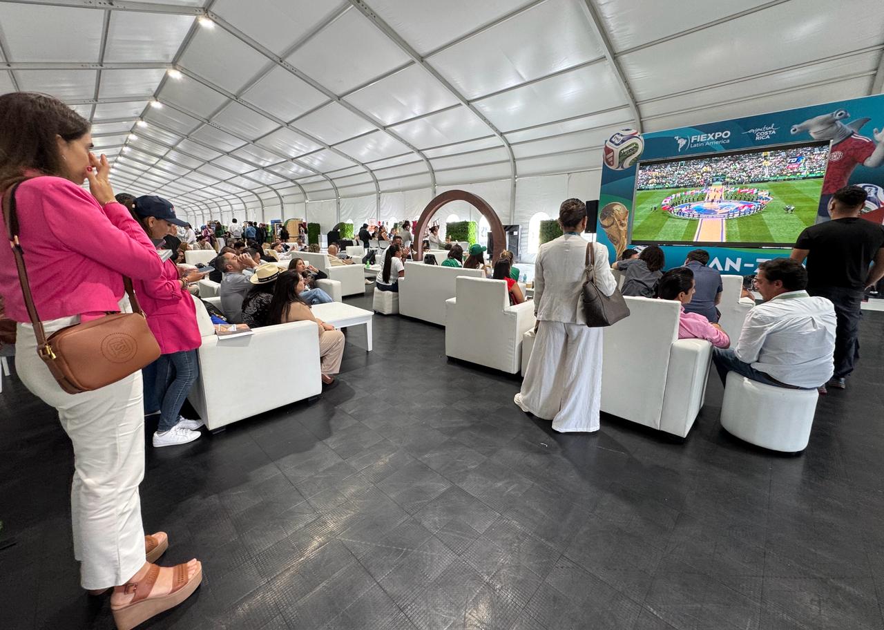 Fotografía En Plano General Que Muestra El Interior De Una Gran Estructura De Carpa Blanca Iluminada Durante El Evento Fiexpo Latin America 2026. En El Espacio, Un Piso Negro Texturizado Sirve De Base Para Múltiples Salas De Estar Compuestas Por Sillones Y Sofás Blancos Donde Los Asistentes Descansan Y Conversan. En El Extremo Derecho, Destaca El Stand Publicitario Azul De Fiexpo Con Una Gran Pantalla Digital Que Transmite Un Evento Deportivo En Un Estadio De Fútbol. En Primer Plano A La Izquierda, Una Mujer Viste Un Saco Rosa Vibrante Y Pantalón Blanco Mientras Observa El Entorno.