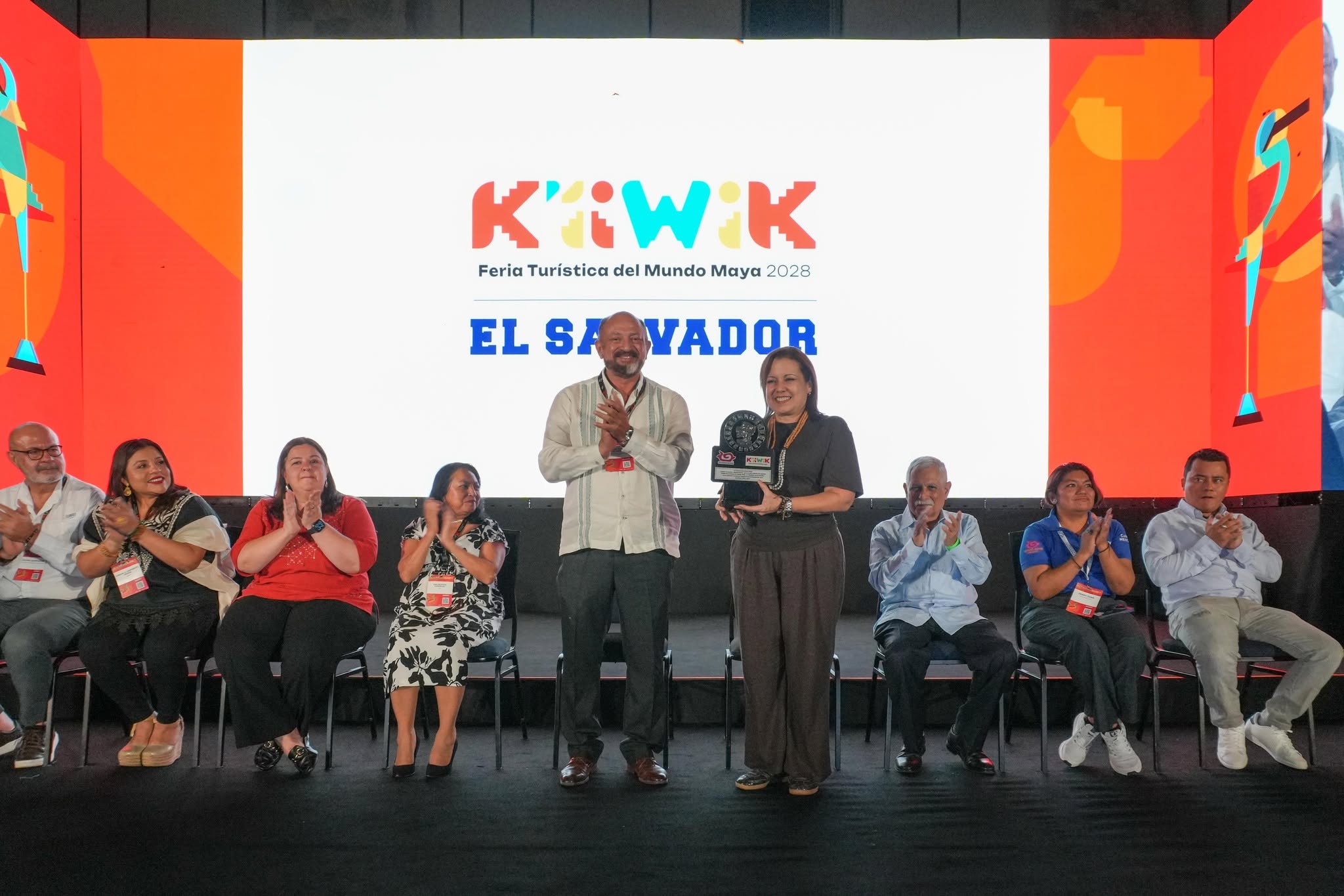 Yucatán consolida con éxito la feria k’íiwik 2026 y sella el futuro de la integración turística en el mundo maya la revista del sureste mx Una Fotografía De Rango Medio Captura A Un Grupo De Personas En Un Escenario Durante Una Presentación Formal, Alineadas Detrás De Una Larga Mesa. En El Centro, Un Hombre Y Una Mujer De Pie Sostienen Un Premio O Un Artefacto Cilíndrico Y Simbólico Sobre Un Pedestal. Una Gran Pantalla Led Detrás De Ellos Muestra El Logotipo &Quot;K'Iiwik&Quot; Y El Texto &Quot;Feria Turística Del Mundo Maya 2028 El Salvador&Quot;. Varias Otras Personas Están Sentadas A Ambos Lados, Aplaudiendo. Las Dos Pantallas Laterales Adyacentes Muestran Gráficos Coloridos De Un Ave O Quetzal Estilizado.