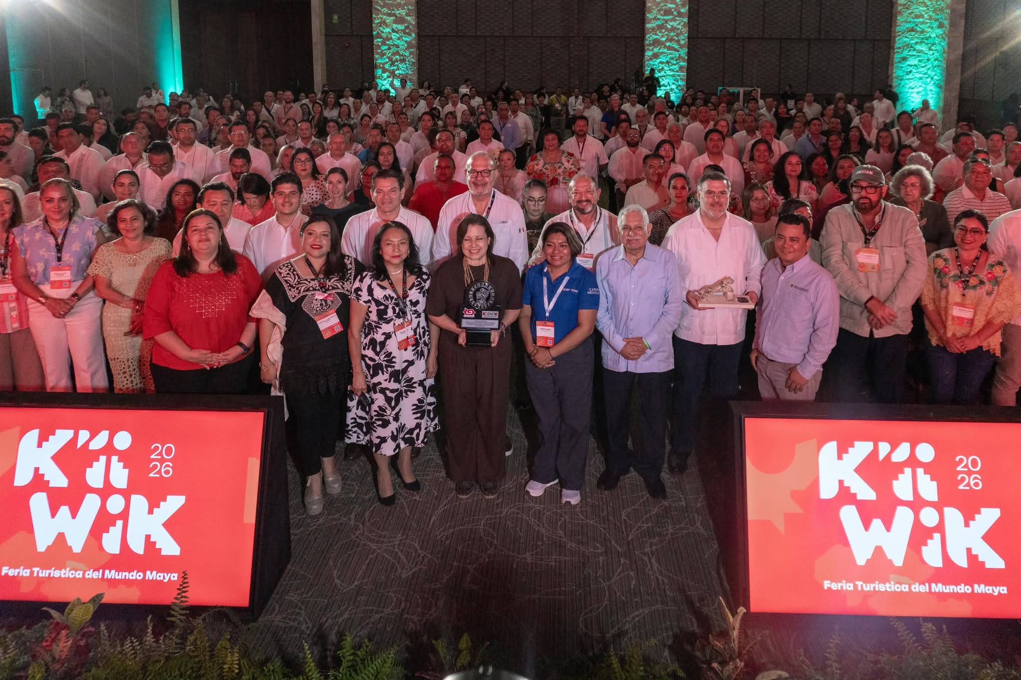 Una Fotografía Grupal A Nivel De Piso En Una Conferencia En Interiores, Con Una Gran Multitud De Asistentes De Pie En El Fondo, Muchos Vestidos De Blanco. En El Centro Del Primer Plano, Un Grupo De Dignatarios Está De Pie, Incluidos Varios Oradores Y Funcionarios, Algunos Sosteniendo Premios O Estafetas. Dos Pantallas Led Brillantes En El Borde Delantero De La Audiencia Muestran El Logotipo De 'K’íiwik 2026' Con El Subtítulo 'Feria Turística Del Mundo Maya'. El Fondo De La Multitud Está Sutilmente Iluminado Con Luces Verdes Y Turquesas.