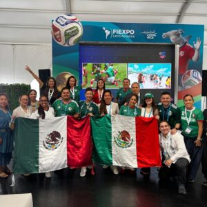 Fotografía Grupal En Plano Entero De Una Delegación Mexicana Posando Sonriente Dentro De Un Recinto De Eventos Durante Fiexpo Latin America 2026 En Costa Rica. El Grupo, Compuesto Por Hombres Y Mujeres, Sostiene Con Orgullo Dos Banderas De México Extendidas Al Frente. Varios Miembros Visten Playeras Verdes De La Selección Mexicana De Fútbol Y Portan Acreditaciones Del Evento Colgadas Al Cuello. Al Fondo Destaca Un Gran Stand Publicitario De Color Azul Con Los Logotipos De Fiexpo Latin America Y &Amp;Quot;Essential Costa Rica&Amp;Quot;, Complementado Con Pantallas Digitales Que Muestran Imágenes Promocionales Y Un Balón De Fútbol Flotante Decorativo.