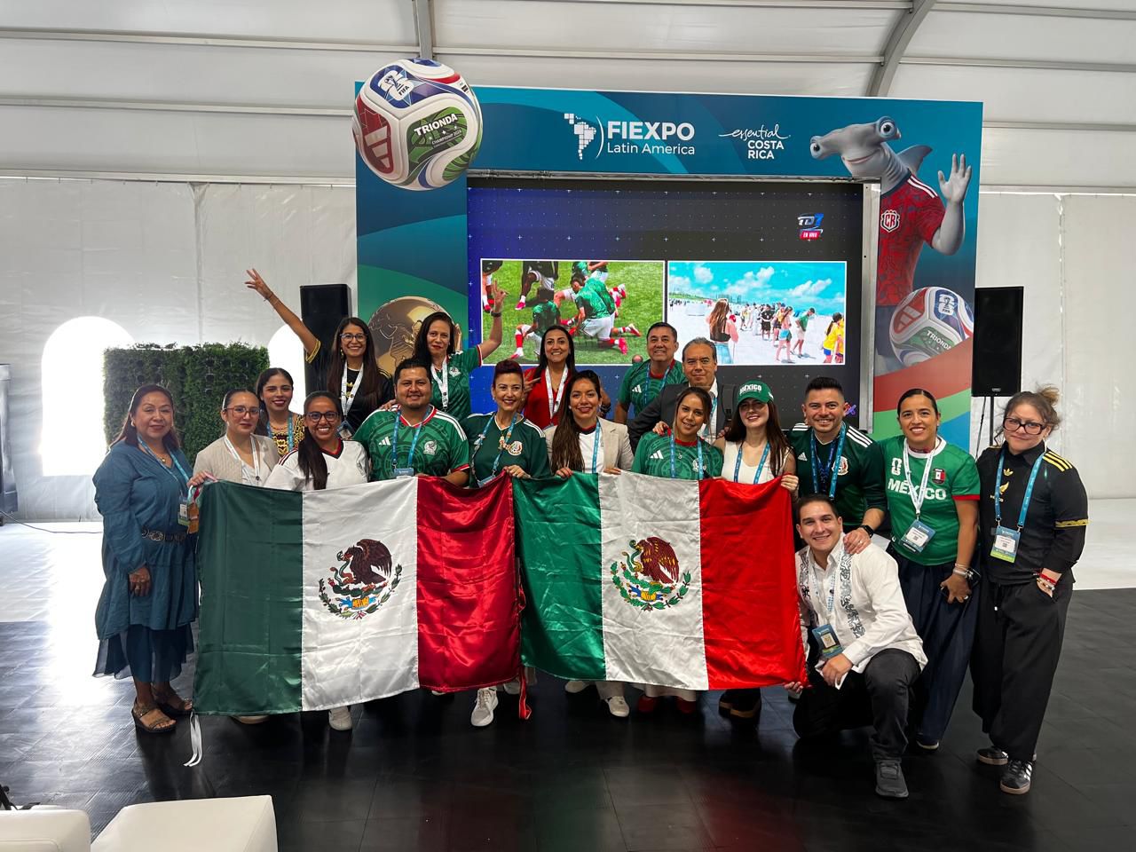 Fotografía Grupal En Plano Entero De Una Delegación Mexicana Posando Sonriente Dentro De Un Recinto De Eventos Durante Fiexpo Latin America 2026 En Costa Rica. El Grupo, Compuesto Por Hombres Y Mujeres, Sostiene Con Orgullo Dos Banderas De México Extendidas Al Frente. Varios Miembros Visten Playeras Verdes De La Selección Mexicana De Fútbol Y Portan Acreditaciones Del Evento Colgadas Al Cuello. Al Fondo Destaca Un Gran Stand Publicitario De Color Azul Con Los Logotipos De Fiexpo Latin America Y &Quot;Essential Costa Rica&Quot;, Complementado Con Pantallas Digitales Que Muestran Imágenes Promocionales Y Un Balón De Fútbol Flotante Decorativo.