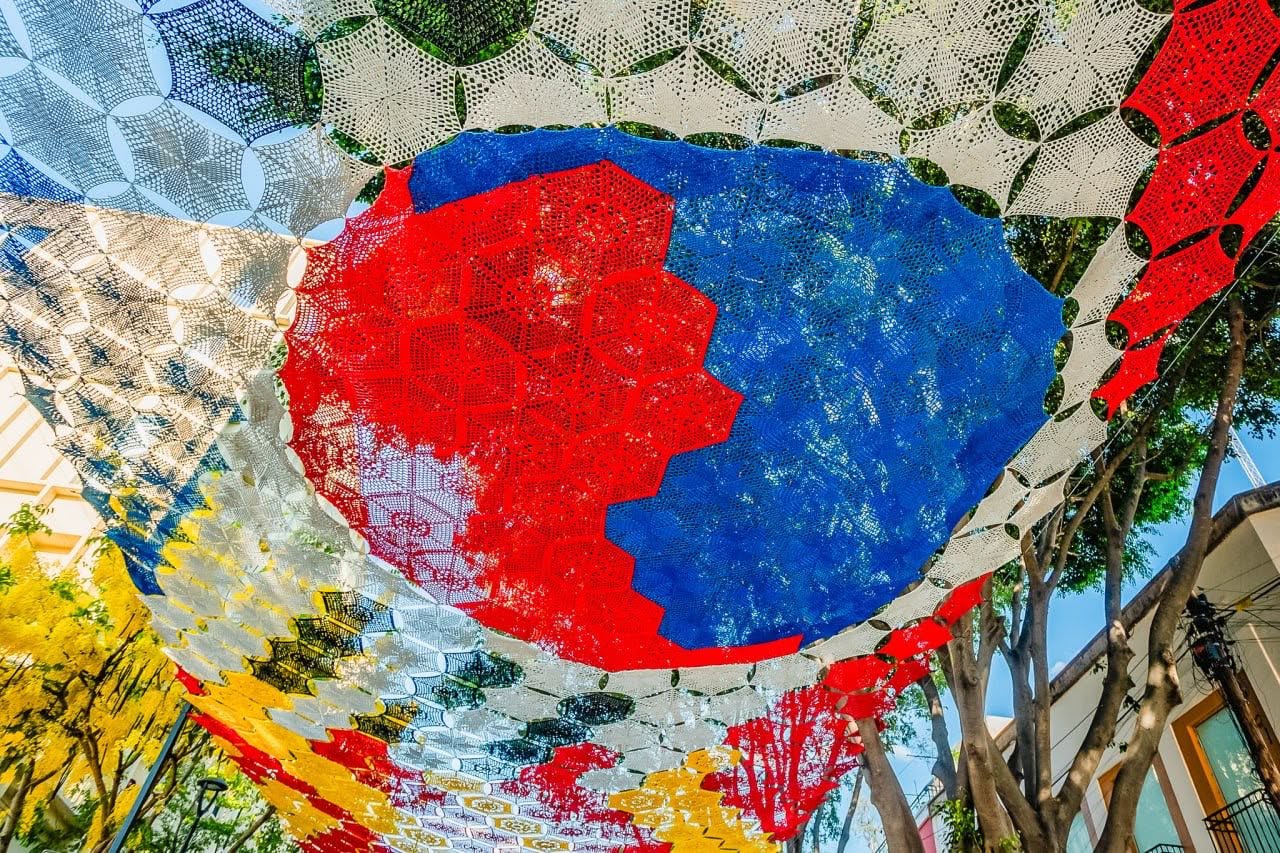 El arte que abraza al fútbol: zapopan inaugura un monumental cielo tejido por manos mexicanas para el mundial 2026 la revista del sureste mx Una Imagen De Plano General En Exteriores Muestra Un Andador Peatonal Moderno Decorado Con Un Monumental &Quot;Cielo Tejido&Quot; Multicolor Suspendido A Lo Largo De La Calle. El Tejido Está Compuesto Por Miles De Hexágonos De Gancho Que Forman Patrones Geométricos, Balones De Fútbol Y Banderas De Distintos Países, Incluyendo México Y Corea Del Sur.