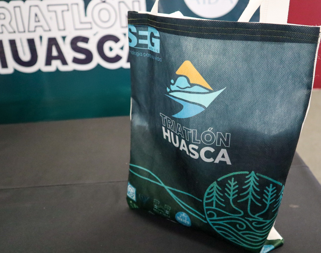 Fotografía En Primer Plano Que Enfoca Una Bolsa Ecológica Promocional De Color Negro Y Verde Oscuro, Colocada De Pie Sobre Una Superficie Plana Oscura. En El Centro De La Bolsa Se Lee Claramente El Texto &Quot;Triatlón Huasca&Quot; En Letras Blancas, Acompañado En La Parte Superior Por Un Logotipo Abstracto Que Simula Montañas Y Olas En Color Amarillo Y Azul, Junto Al Logotipo De Seg México.
