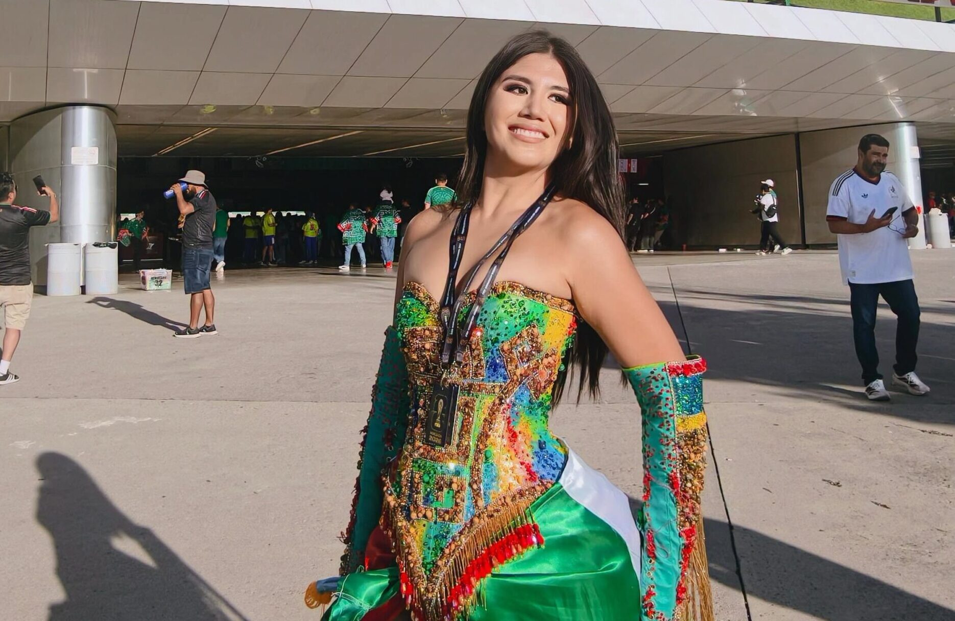 La Influencer Friday Frida Sonríe Posando Frente Al Acceso Principal Del Estadio Guadalajara (Estadio Akron). Viste Un Elaborado Traje Artesanal Strapless Multicolor, Con Pedrería Brillante En Tonos Verdes, Dorados Y Rojos, Manguillas Decoradas Con Flecos Dorados A Juego, Y Una Falda Satinada Con Los Colores De La Bandera Mexicana Verde Y Rojo.