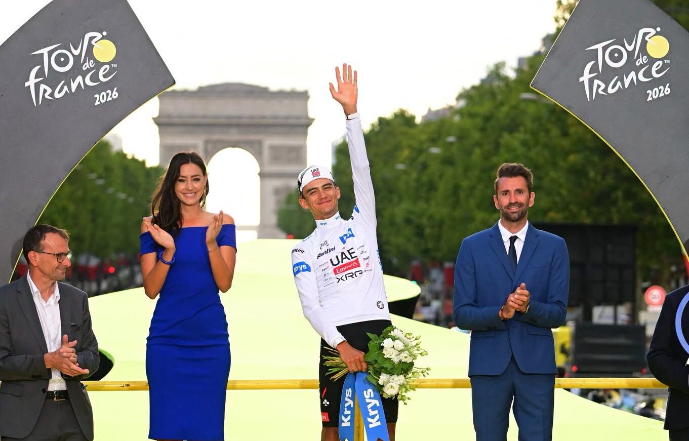 El Ciclista Mexicano Isaac Del Toro Saluda Desde El Podio Vistiendo El Maillot Blanco Como Campeón Joven Del Tour De Francia 2026, Con El Arco De Triunfo De París Al Fondo.