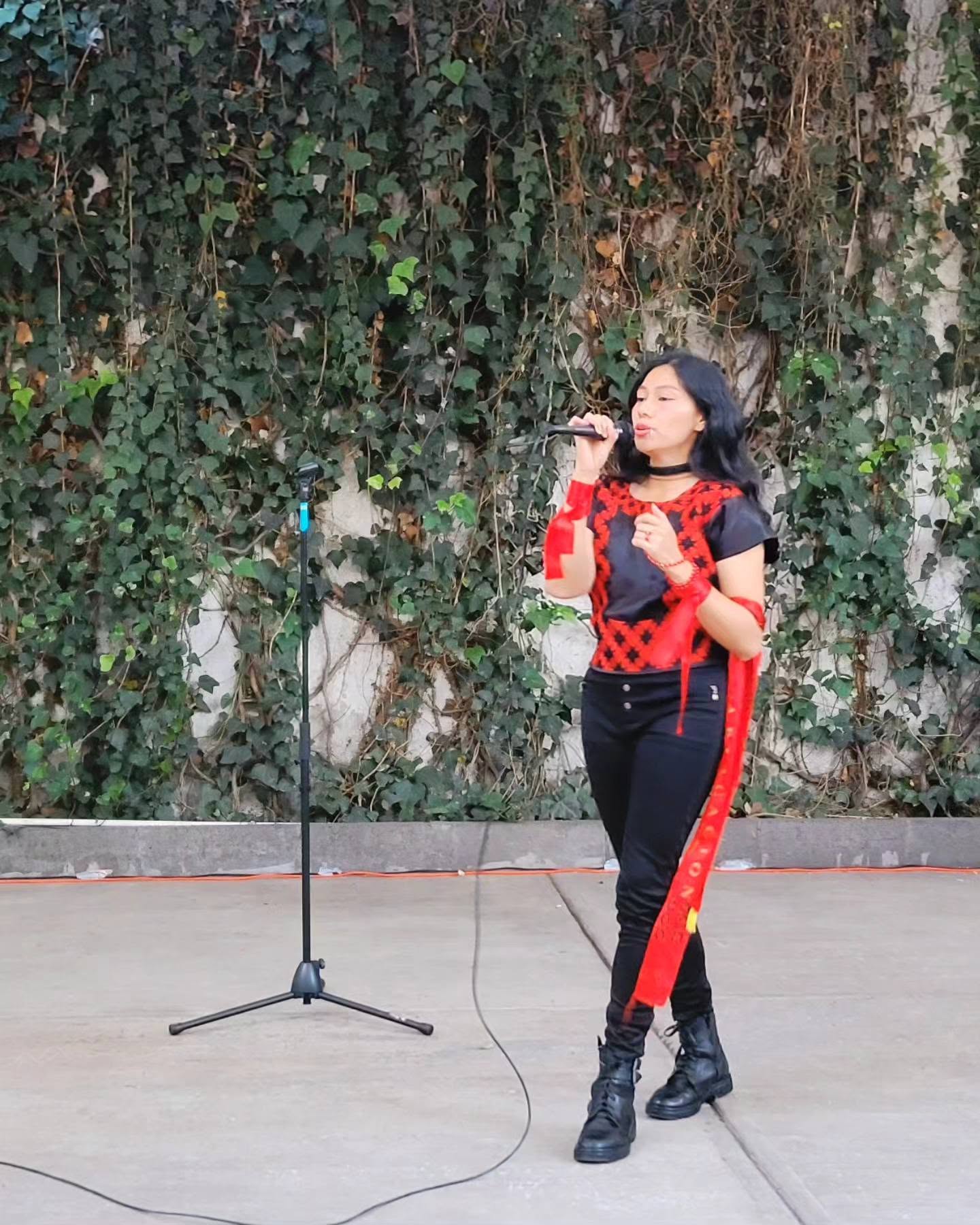 Azalea báalam: la voz mexicana que revitaliza el maya y el náhuatl a ritmo de pop la revista del sureste mx Azalea Báalam Cantando Con Un Micrófono Frente A Un Muro Cubierto De Hiedra, Vistiendo Ropa Negra Con Bordados Rojos Tradicionales.
