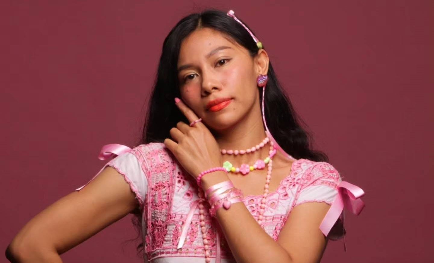 : Retrato De La Cantautora Mexicana Azalea Báalam Vistiendo Una Blusa Blanca Con Bordados Y Accesorios Rosas, Sobre Un Fondo Color Vino.