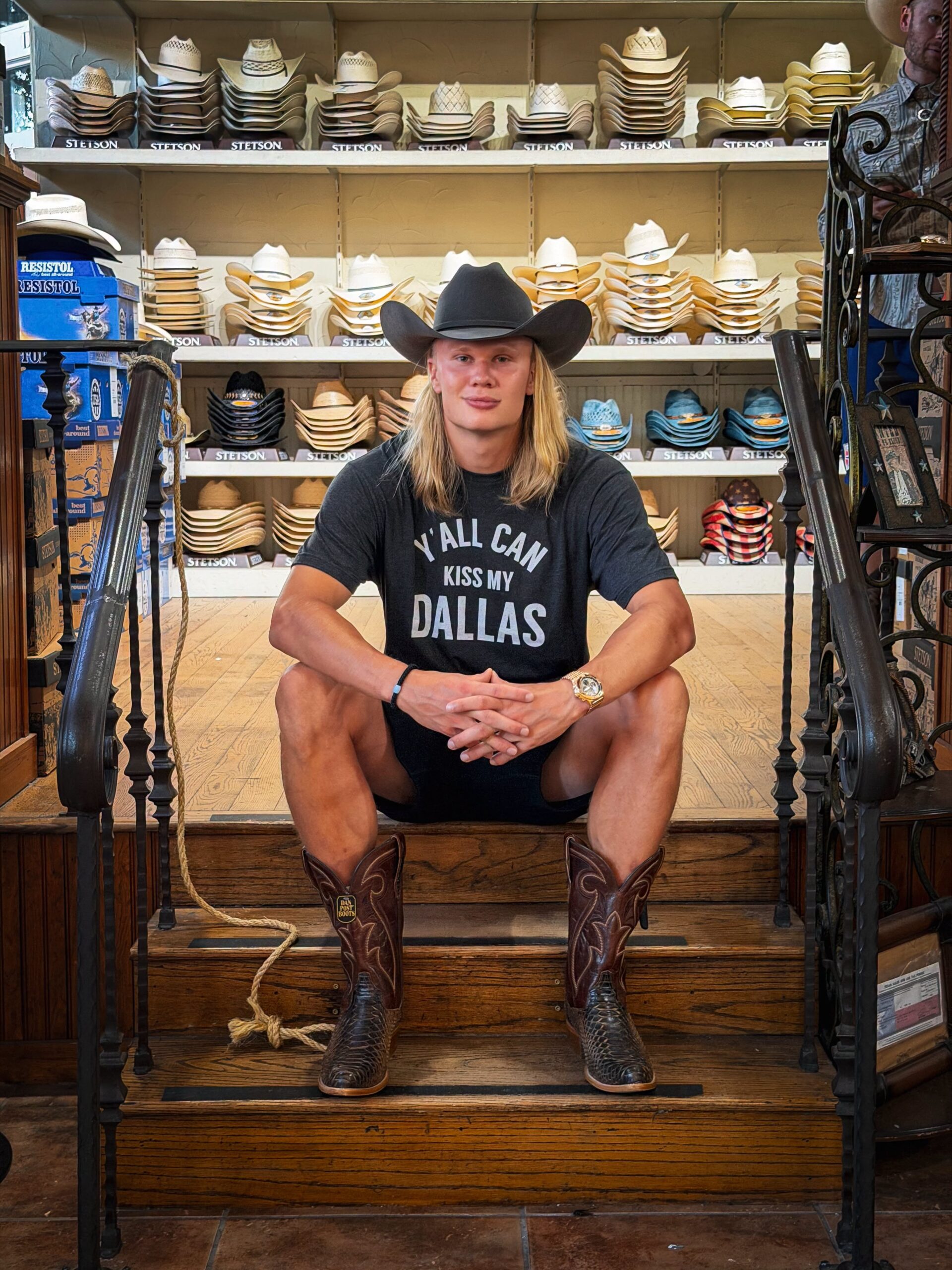 Erling haaland responde a méxico tras festejo vikingo en mundial 2026 la revista del sureste mx Erling Haaland Sentado En Una Escalera De Madera Dentro De Una Tienda De Sombreros Vaqueros, Vistiendo Botas, Sombrero Negro Y Una Camiseta Con El Lema &Quot;Y'All Can Kiss My Dallas&Quot;.