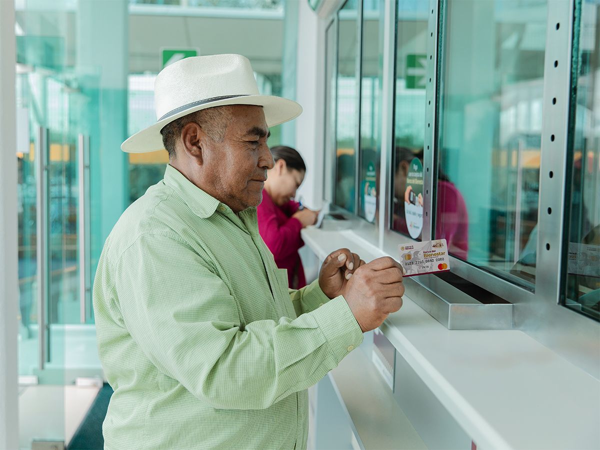 Un Hombre Mayor Con Sombrero Y Camisa Verde Sostiene Su Tarjeta Del Bienestar Frente A La Ventanilla De Atención En Una Sucursal Bancaria.
