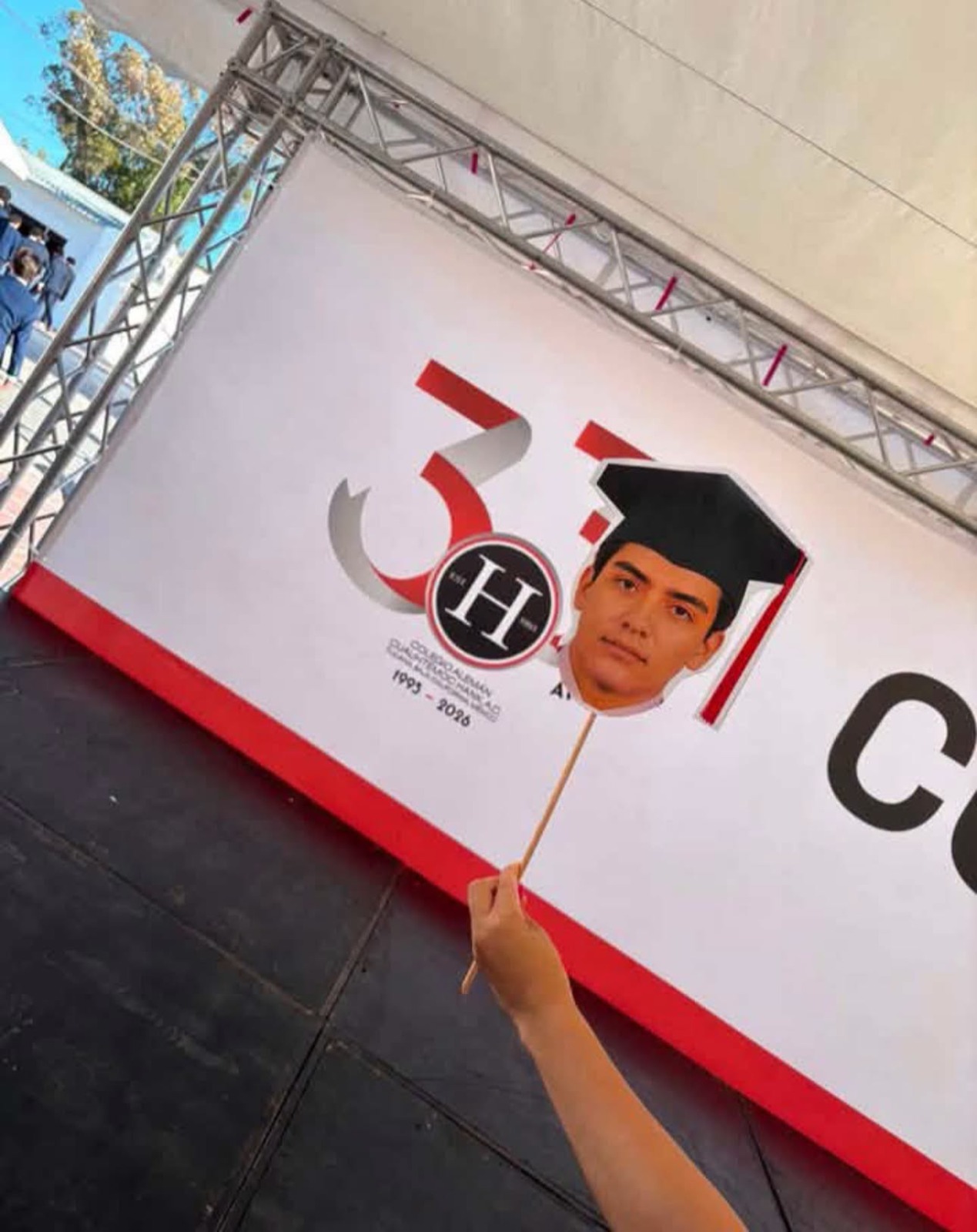 Gilberto mora: entre el éxito mundialista y su emotiva graduación de preparatoria la revista del sureste mx Una Mano Sostiene Un Recorte De Cartón Con La Cara De Gilberto Mora Y Un Birrete De Graduación Frente A Un Gran Cartel Del Evento De Graduación Del Colegio Alemán Cuauhtémoc Hank.