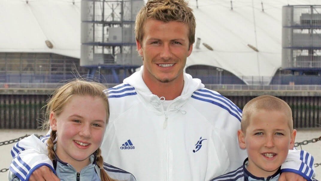 Fotografía De Archivo Que Muestra A Un Joven Harry Kane Y A Katie Goodland (Su Futura Esposa) Cuando Eran Niños, Posando Sonrientes Junto A David Beckham En La Inauguración De Su Academia De Fútbol En 2005. Los Tres Llevan Chaquetas Deportivas Grises De La Academia.