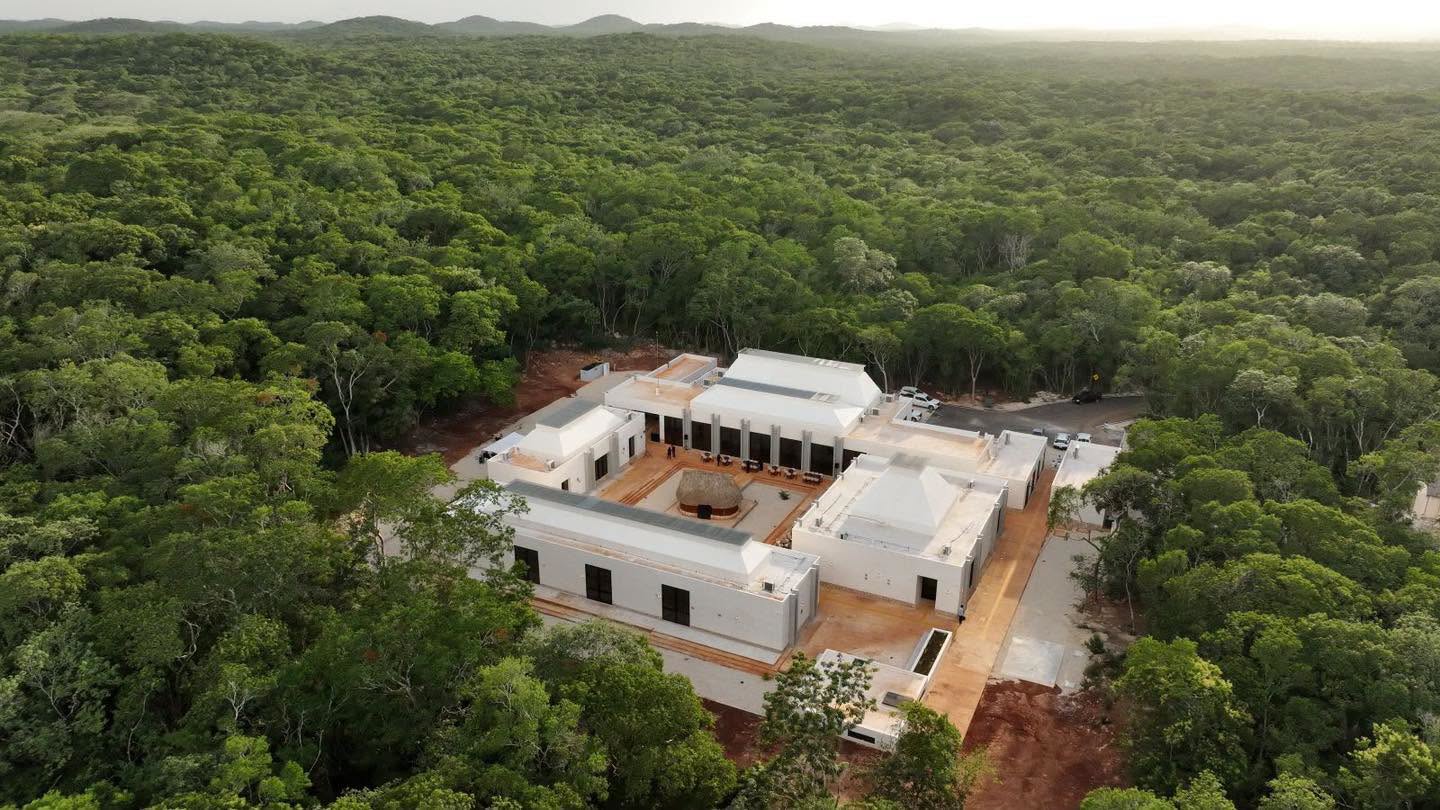 Inauguran dos nuevos museos para fortalecer el legado de la cultura maya en yucatán la revista del sureste mx Vista Aérea Del Museo Arqueológico Del Puuc En Kabah. Un Moderno Complejo Blanco De Varios Edificios, Con Cubiertas Piramidales Bajas Y Un Patio Central Que Rodea Una Gran Estructura De Techo De Paja, Está Enclavado En Medio De Una Densa Selva De Copas De Árboles De Color Verde Intenso Bajo Un Cielo Brumoso. El Sol Incide De Frente En Las Fachadas, Realzando Sus Formas Geométricas. Un Camino De Grava Se Extiende Hasta El Complejo.