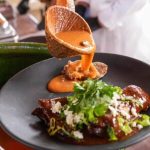 Oaxaca enaltece su riqueza gastronómica con el festival de los moles 2026 la revista del sureste mx Oaxaca Enaltece Su Riqueza Gastronómica Con El Festival De Los Moles 2026 — Cucharón De Mole Artesanal Vertiendo Una Espesa Salsa De Mole Sobre Un Platillo Tradicional En Plato Negro, Decorado Con Queso Fresco Y Cilantro Durante La Presentación Gastronómica.
