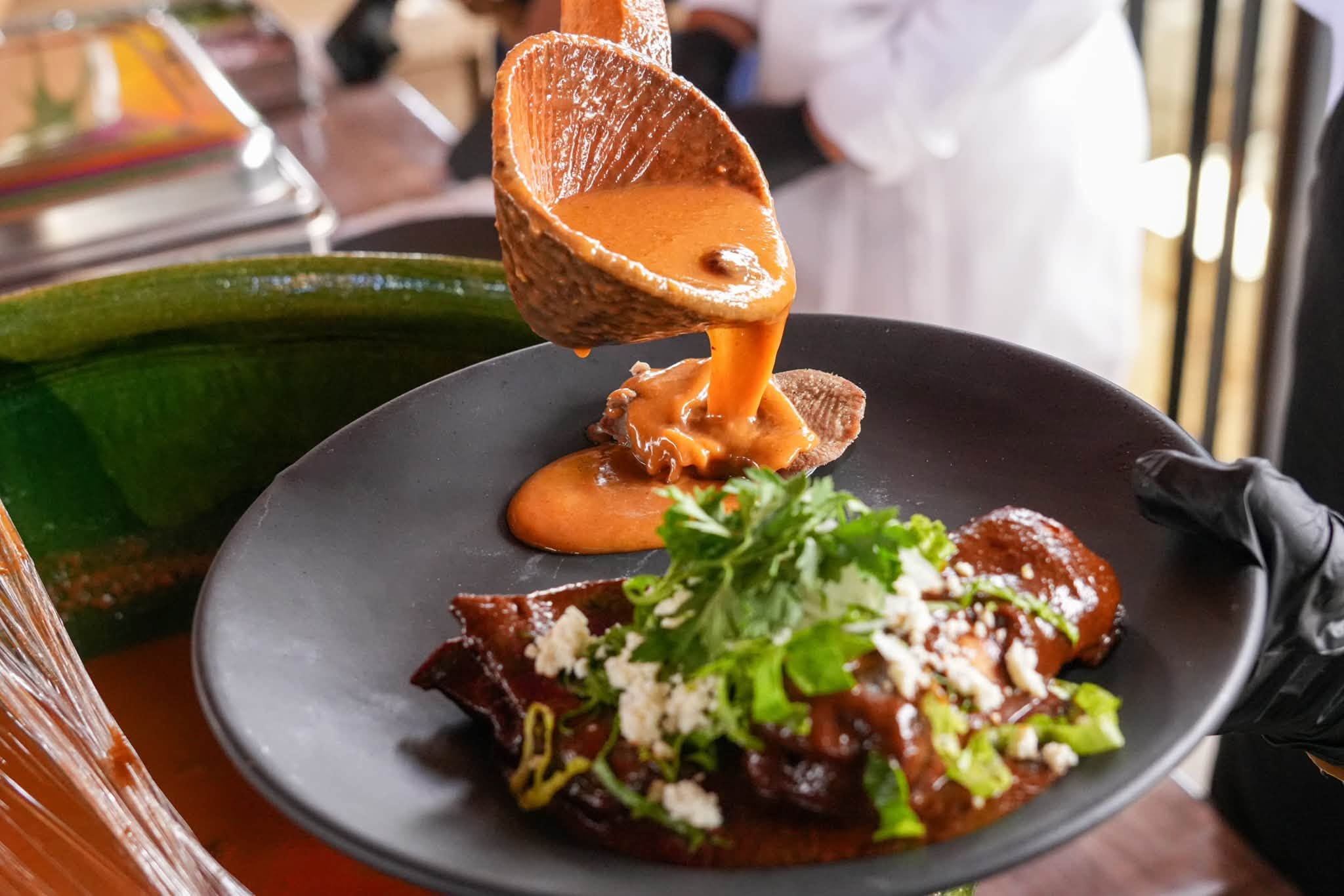 Oaxaca Enaltece Su Riqueza Gastronómica Con El Festival De Los Moles 2026 — Cucharón De Mole Artesanal Vertiendo Una Espesa Salsa De Mole Sobre Un Platillo Tradicional En Plato Negro, Decorado Con Queso Fresco Y Cilantro Durante La Presentación Gastronómica.