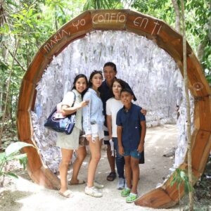 Una Familia Sonriente Posa En El Santuario De María Desatadora De Nudos, Ejemplo Del Crecimiento Del Turismo Religioso En Cancún. El Arco De Madera Tiene La Leyenda «María, Yo Confío En Ti» Y Está Decorado Con Listones De Peticiones De Peregrinos.