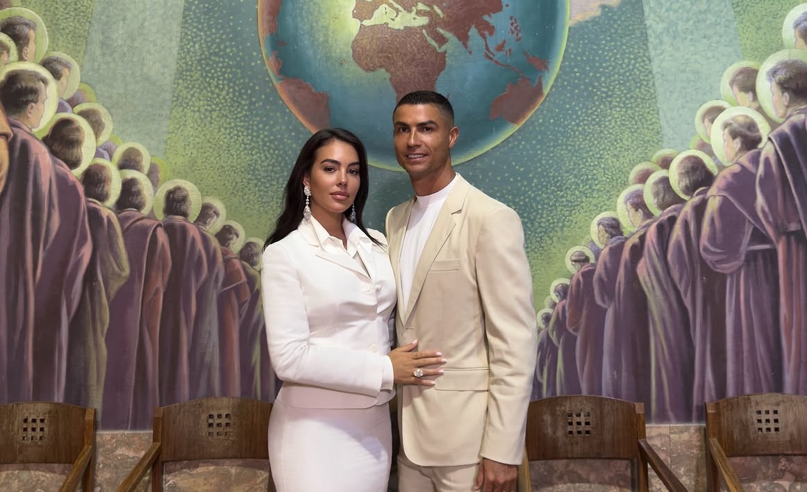 Amor, lujo y un acuerdo millonario: todos los detalles de la íntima boda civil de cristiano ronaldo y georgina rodríguez la revista del sureste mx Cristiano Ronaldo Y Georgina Rodríguez Posan Elegantemente Frente A Un Mural Religioso Detallado. Georgina Lleva Un Traje De Falda Blanco De Gucci Y Cristiano Un Traje Beige De Gucci, Con Joyas De Chopard De 75 Millones De Dólares De La Colección Garden Of Kalahari De Vogue. El Mural Muestra Un Mapa Del Mundo Y Figuras De Santos.