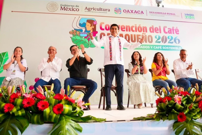 El Gobernador De Oaxaca, Salomón Jara Cruz, De Pie En El Centro De Un Escenario Presídium Con Los Brazos Abiertos En Gesto De Bienvenida Durante La Inauguración De La Convención Del Café Oaxaqueño 2026. A Su Lado Se Encuentran Sentados Funcionarios Estatales, Incluida La Presidenta Del Dif Estatal, Irma Bolaños Quijano, Aplaudiendo. En El Fondo Se Aprecia La Lona Oficial Con Los Logotipos Del Gobierno De México, La Secretaría De Agricultura, El Gobierno De Oaxaca Y La Sefader, Mientras Que En Primer Plano Destacan Arreglos Florales Coloridos Con Gerberas Rojas Y Lirios.