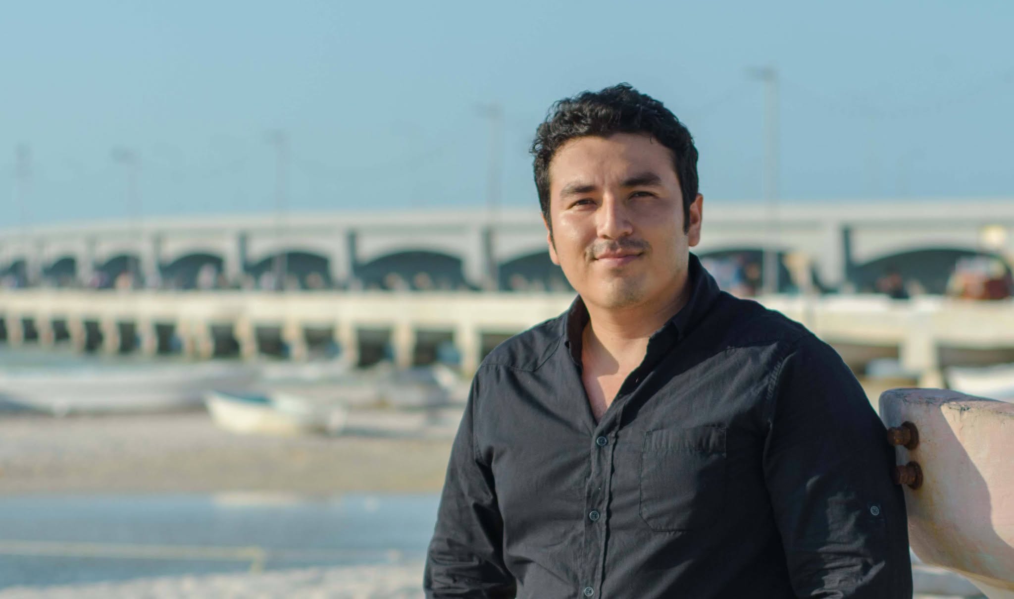 Un Retrato En Plano Medio Del Cineasta Yucateco Víctor Rejón, Codirector De &Quot;Las Voces Del Despeñadero&Quot;, De Pie En Una Playa Cerca Del Mar, Con El Muelle De Progreso Al Fondo Bajo Un Cielo Despejado. Él Lleva Una Camisa Negra.