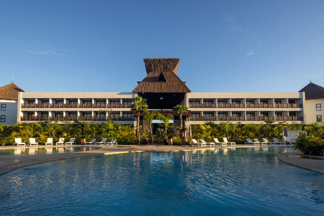 Una Vista Frontal Y Simétrica Del Hotel Mundo Maya Desde El Borde De Su Piscina Azul. El Hotel Es Un Edificio Moderno De Tres Pisos Con Balcones De Madera, Coronado Por Una Gran Estructura Central De Palapa De Techo De Paja. Un Jardín Tropical Y Hileras De Tumbonas Separan El Edificio De La Piscina Bajo Un Cielo Azul Despejado Con Luz Solar Cálida.