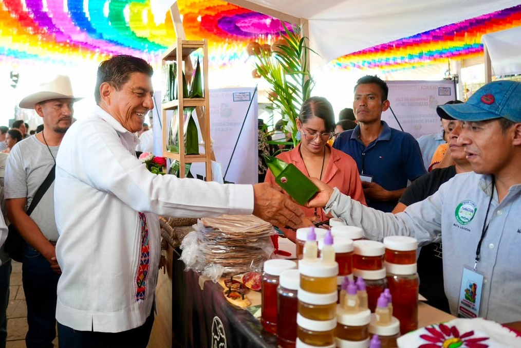 El Gobernador De Oaxaca, Salomón Jara Cruz, Vistiendo Una Guayabera Blanca Con Bordados Artesanales Coloridos, Sonríe Mientras Saluda A Un Productor En Un Stand De La Convención Del Café Oaxaqueño 2026. El Puesto Exhibe Empaques Verdes De Café, Frascos De Miel Y Productos Locales Bajo Un Techo Decorado Con Papel Picado De Múltiples Colores.