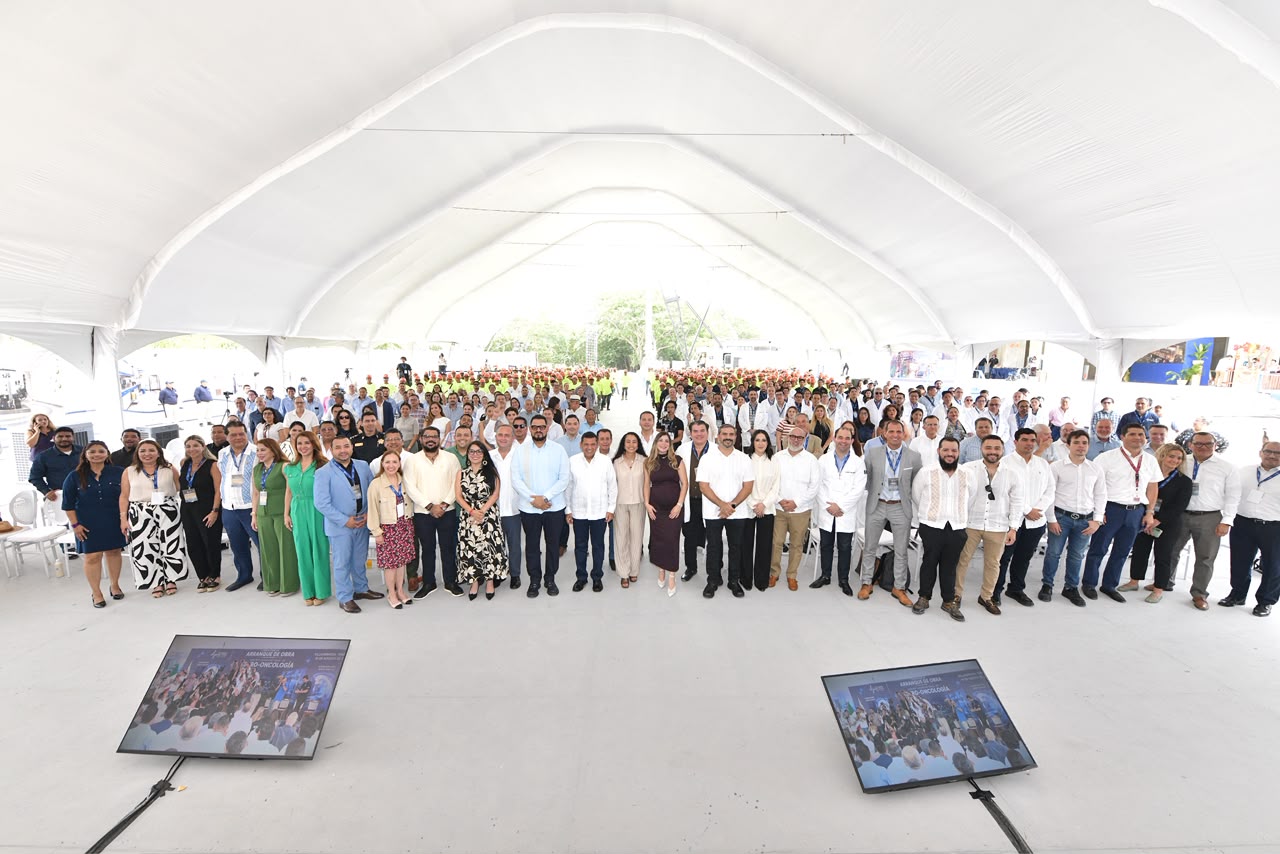 Una Toma Elevada Y De Gran Angular Muestra A Un Grupo Numeroso De Personas Posando Para Una Fotografía De Grupo En Un Gran Lugar Cubierto Para Eventos, Posiblemente Una Carpa Para Eventos. Se Distinguen Varias Filas De Personas, Y Los Invitados De Honor Están De Pie En La Fila Delantera. Dos Monitores Que Transmiten Imágenes En Vivo Están En El Suelo En Primer Plano.