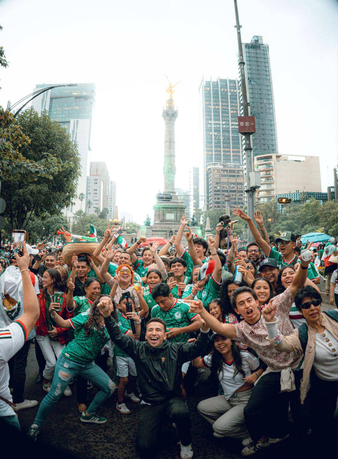 La copa del mundo 2026 catapulta el turismo internacional en méxico: el impacto histórico durante su primer mes la revista del sureste mx Una Fotografía Vertical De Plano Medio Que Captura A Una Multitud Exultante De Aficionados De La Selección Nacional De Fútbol De México Celebrando En Las Calles De La Ciudad De México. El Punto Focal Es Un Joven Sonriente En Primer Plano Que Se Arrodilla Con Los Brazos En Alto. Detrás De Él, Una Multitud De Hombres Y Mujeres Llenan La Calle, Vistiendo Camisetas Verdes De La Selección De México. Levantan Las Manos En El Aire, Animando Y Cantando, Y Algunos Sostienen Banderas Mexicanas Pequeñas. Al Fondo, Se Elevan Modernos Edificios De Oficinas De Vidrio Y El Icónico Monumento Del Ángel De La Independencia, Adornado Con Su Estatua Dorada. La Luz De La Tarde Es Suave Y El Ambiente Es Vibrante Y Lleno De Energía. La Imagen Está Tomada Desde Una Perspectiva Ligeramente Baja.