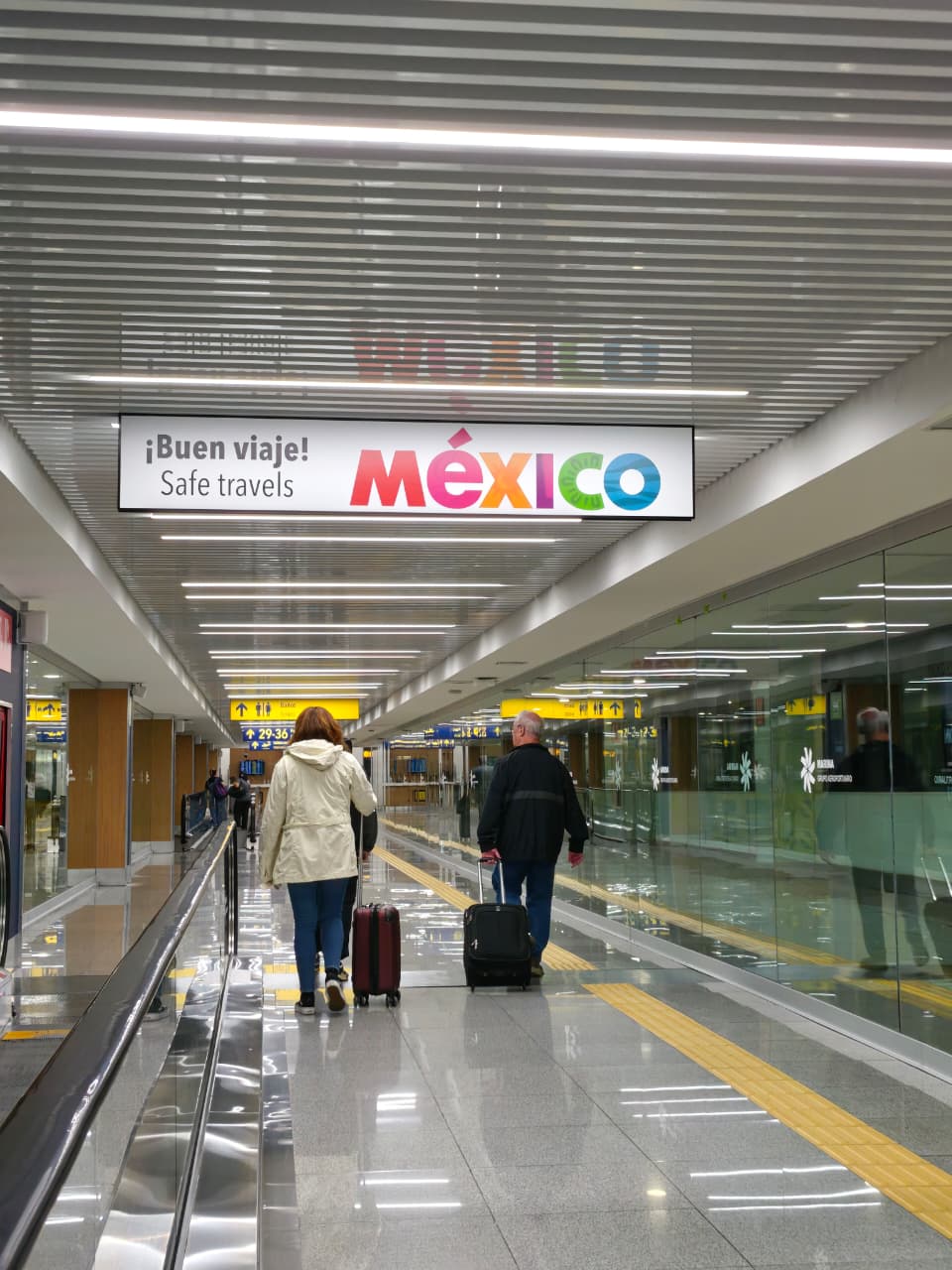 Una Fotografía De Ángulo Medio Y Perspectiva Profunda Captura El Interior De Un Moderno Y Limpio Pasillo De La Terminal De Un Aeropuerto. En Primer Plano, Dos Viajeros Caminan Alejándose De La Cámara Sobre Una Cinta Transportadora Móvil. La Persona De La Izquierda Tiene Cabello Castaño Corto Y Viste Una Chaqueta Blanca Holgada, Jeans Azules Y Zapatillas Oscuras, Y Arrastra Una Maleta De Mano De Color Rojo Oscuro.