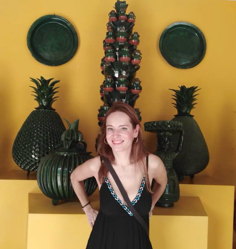 La Actriz Gaby Platas Sonríe Posando Frente A Una Pared Amarilla Decorada Con Artesanías Mexicanas De Cerámica En Vidriado Verde, Incluyendo Esculturas En Forma De Piñas Y Vasijas Tradicionales.