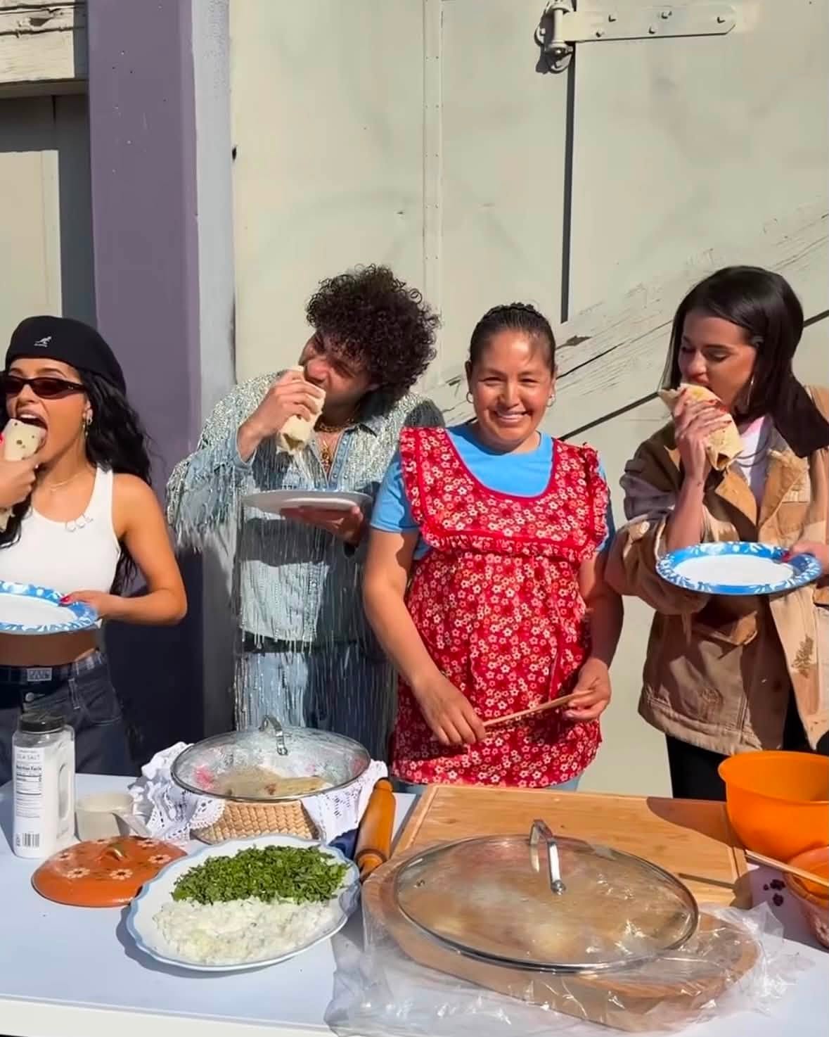 Fotografía En Exterior Donde Selena Gomez, Benny Blanco Y Becky G Muerden Al Mismo Tiempo Un Burrito Tradicional Mexicano. En El Centro De La Imagen, La Creadora De Contenido Rebeca Coss Sonríe A La Cámara Vistiendo Una Playera Azul Y Un Mandil Rojo Con Estampado Floral. A La Derecha, Selena Gomez Inclina La Cabeza Para Dar Un Bocado A Su Burrito Sobre Un Plato Desechable. A La Izquierda, Benny Blanco, Con Una Chaqueta De Mezclilla Decorada Con Flecos Plateados, Y Becky G, Vistiendo Un Top Blanco, Gorra Negra Y Gafas Oscuras, Disfrutan También De Sus Alimentos. En Primer Plano Se Observa Una Mesa Con Guarniciones Como Cilantro, Cebolla, Salsas Y Recipientes Con Tortillas De Harina Caseras.