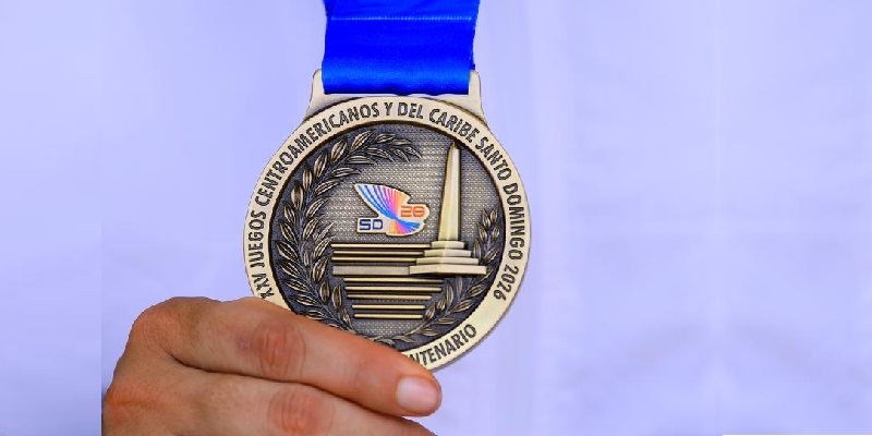 México rompe su récord absoluto en los centroamericanos: suma 146 oros en santo domingo 2026 la revista del sureste mx Primer Plano De Una Mano Sosteniendo Una Medalla De Bronce Oficial Con Cinta Azul, Que Muestra El Relieve Del Monumento De Santiago Y El Logotipo De Los Juegos Centroamericanos Y Del Caribe Santo Domingo 2026.