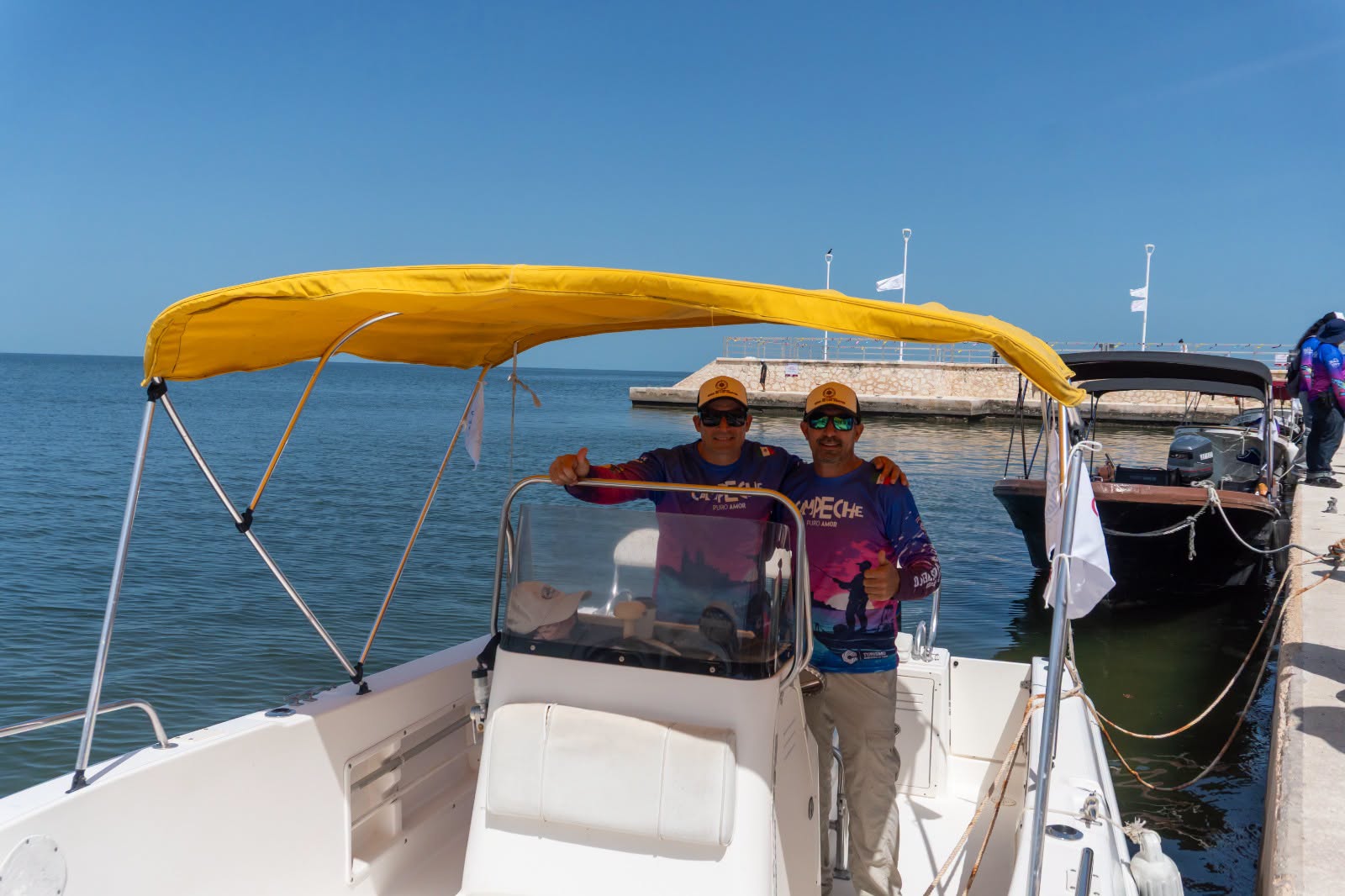 Descubre campeche desde el mar: navega la bahía y los manglares en el muelle ‘rosa de los vientos’ la revista del sureste mx Dos Hombres Con Camisetas Técnicas De Campeche Posan Sonriendo Y Con El Pulgar Hacia Arriba A Bordo De Una Lancha Blanca Con Toldo Amarillo, Atracada En Un Muelle Bajo Un Cielo Azul Y Despejado.