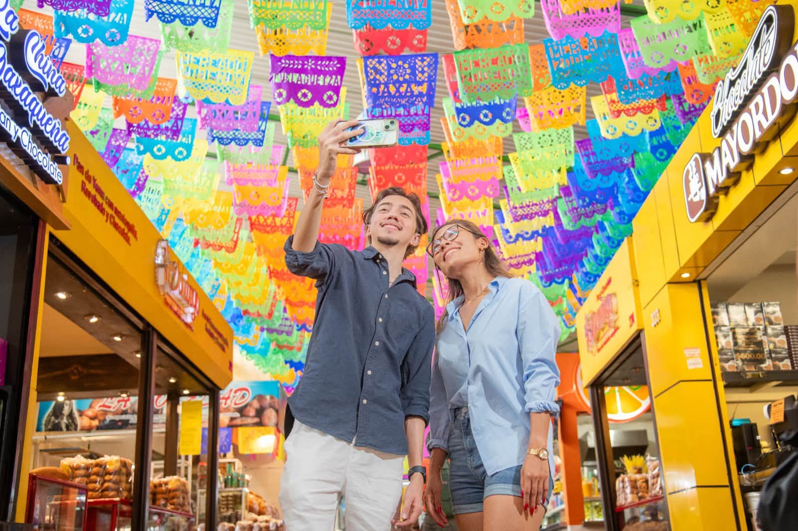 Oaxaca conquista el ranking global: la capital oaxaqueña se posiciona entre los destinos turísticos más felices del mundo la revista del sureste mx Pareja De Turistas Sonriendo Y Tomándose Una Selfie Bajo Colorido Papel Picado En Un Mercado De Oaxaca.