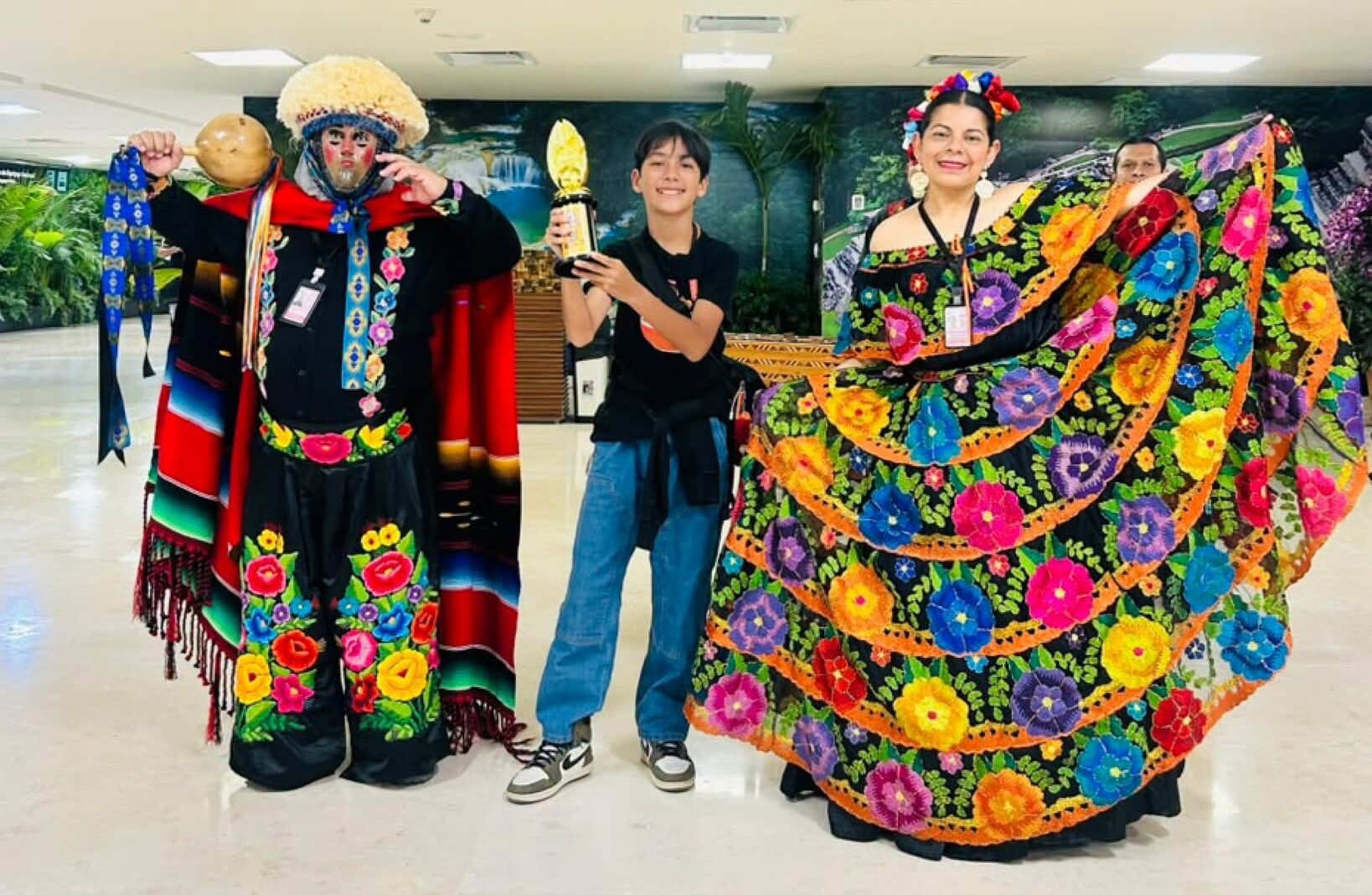 Niño Chiapaneco Sonriendo Mientras Sostiene Un Trofeo Del Campeonato Internacional Aloha Mental Arithmetic, Acompañado Por Personas Vestidas Con Trajes Tradicionales De Parachico Y Chiapaneca.